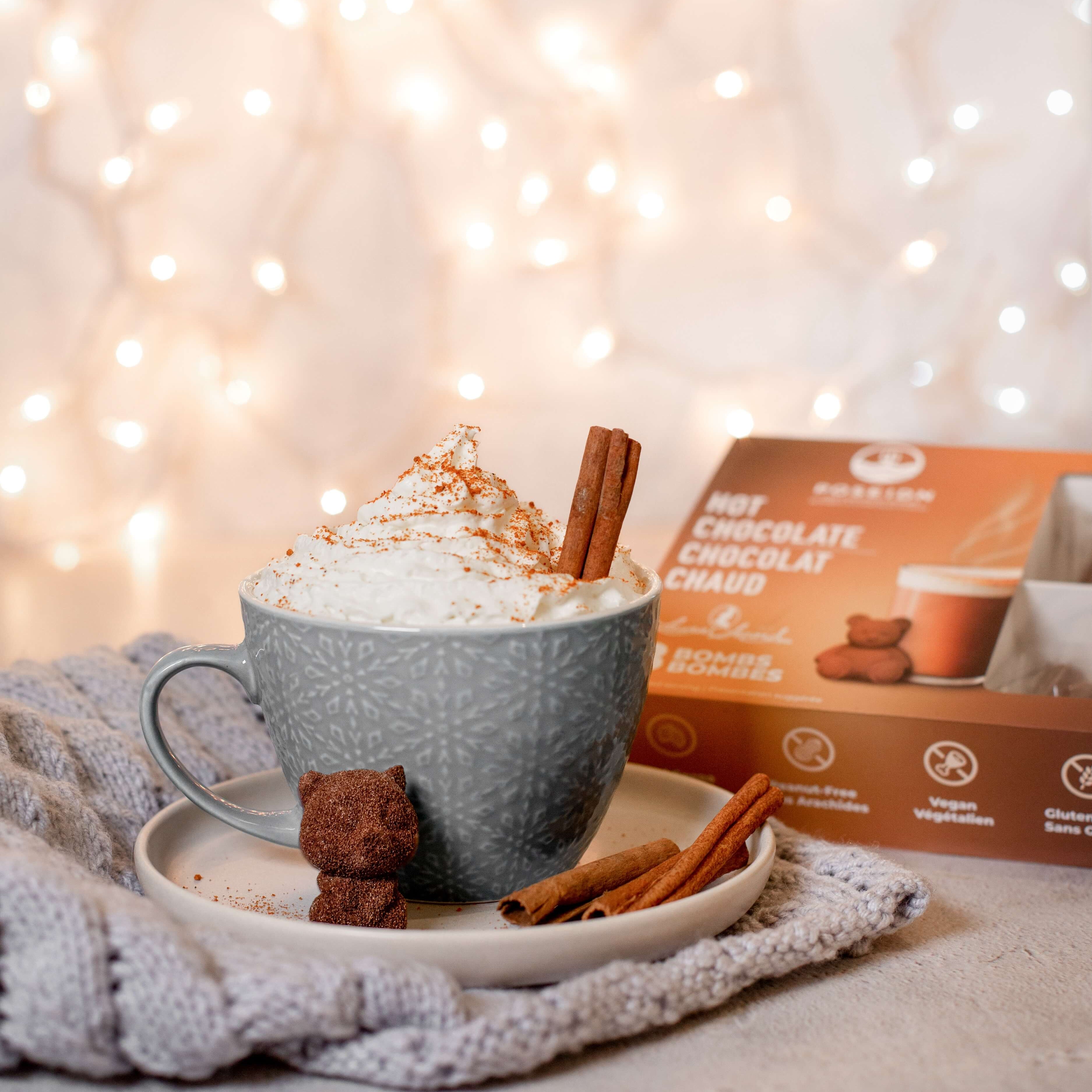 Hot chocolate with whipped cream, a cinnamon stick, and a small bear-shaped cookie on a plate, with a box of hot chocolate in the background.