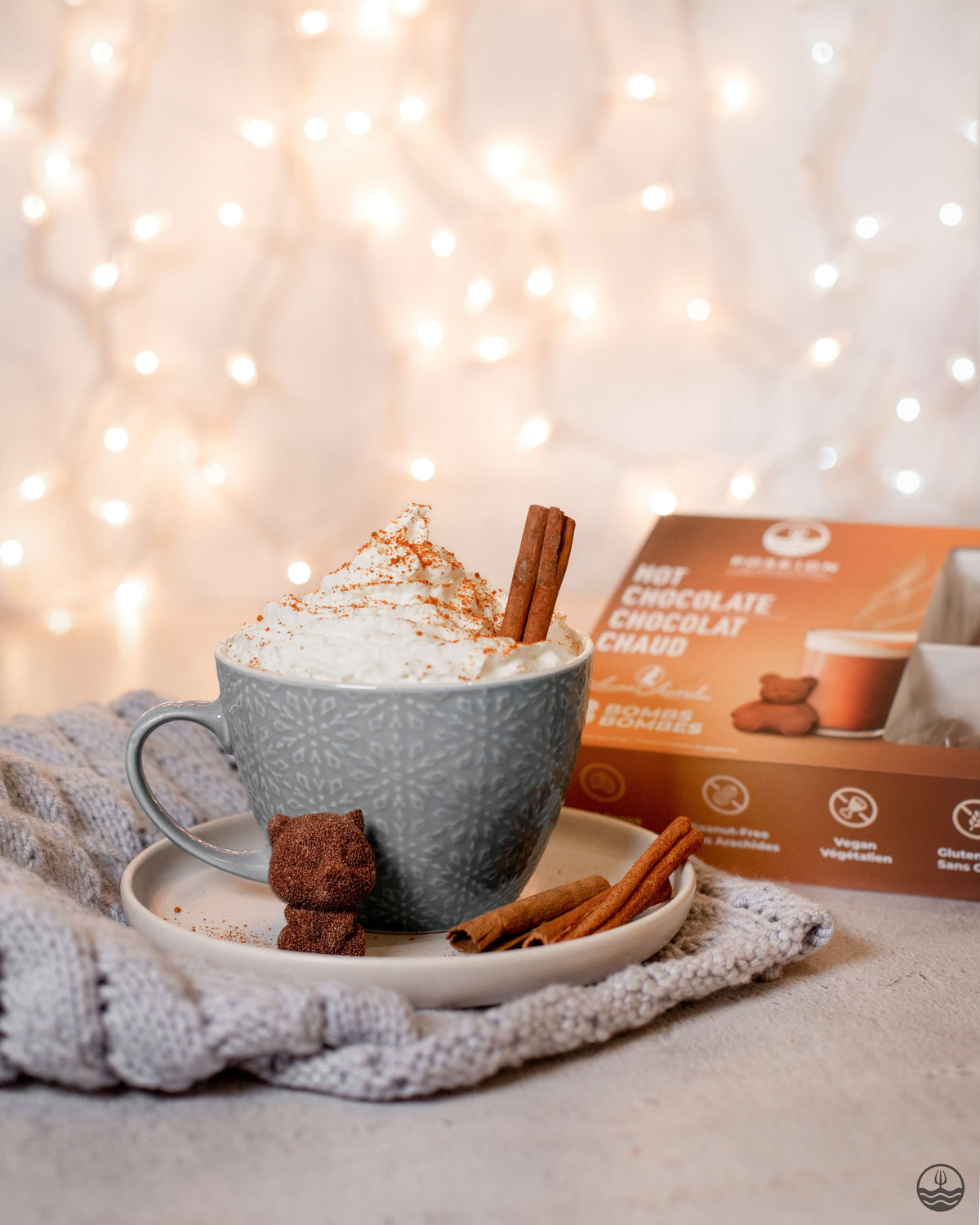 Hot chocolate with whipped cream, a cinnamon stick, and a small bear-shaped cookie on a plate, with a box of hot chocolate in the background.