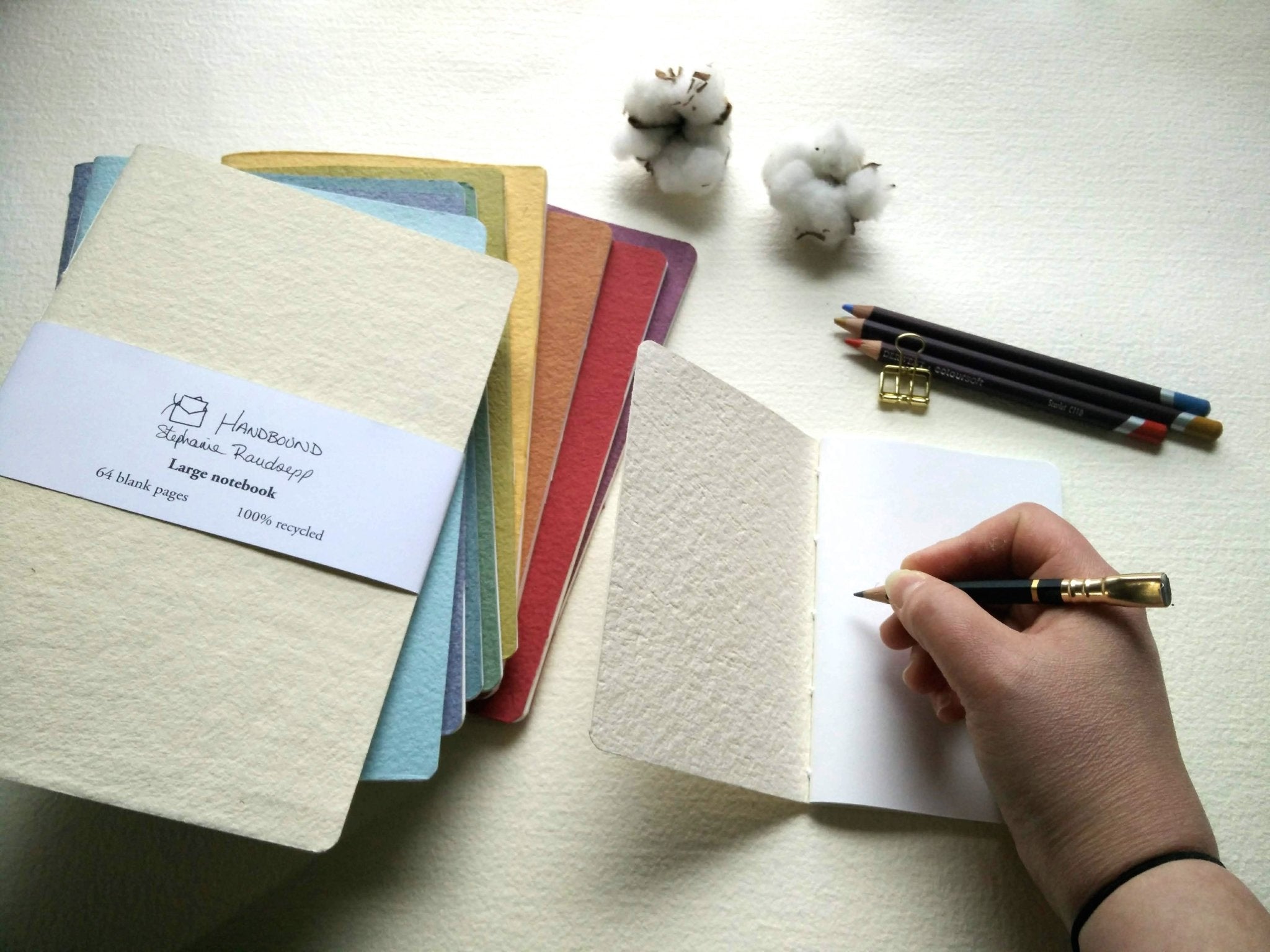 Color swatch book with notebooks and a hand holding a pen on a hand stitched plantable paper notebook on a light grey background Creative Artifacts HQ