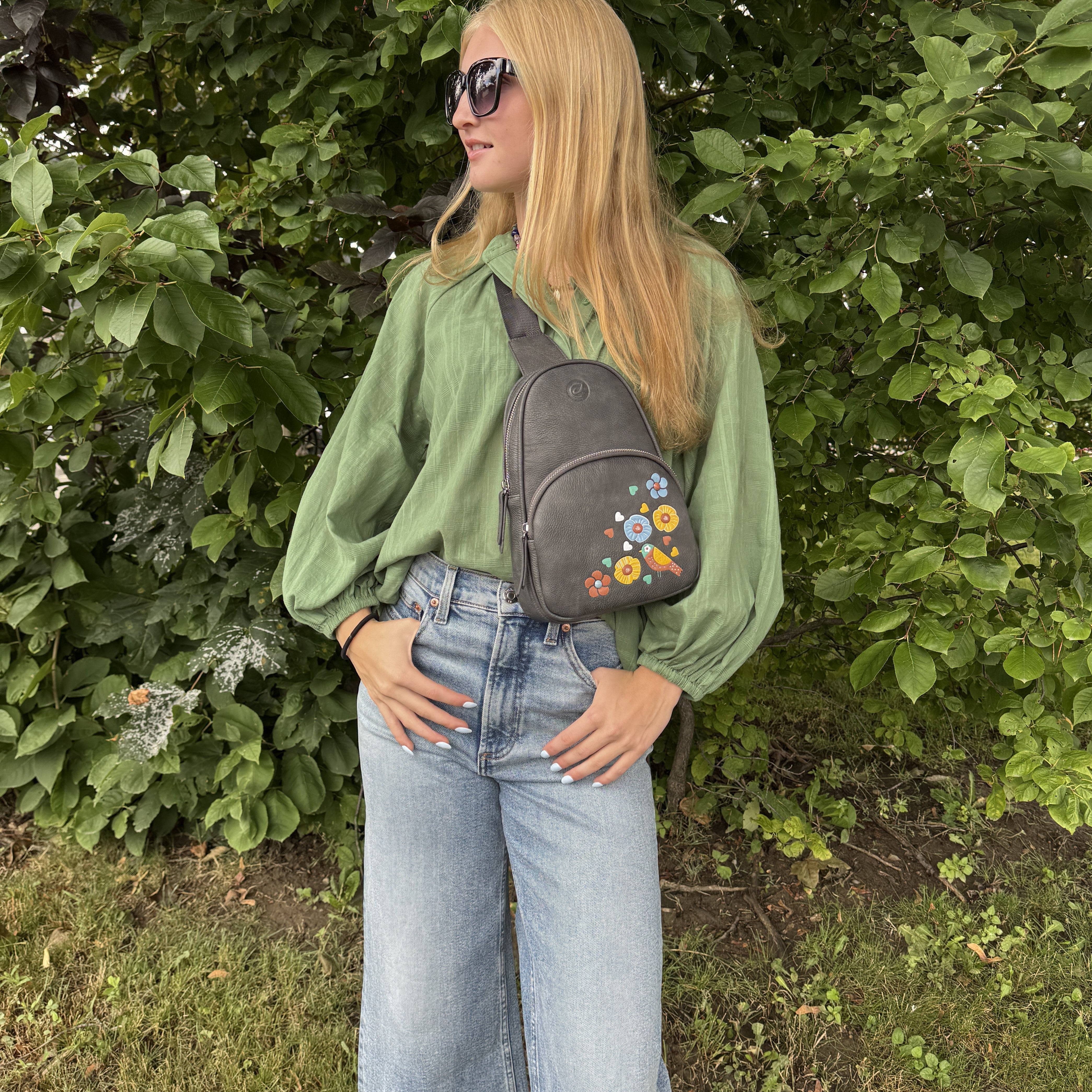 Woman wearing a green blouse and blue jeans with a floral gray bag, standing against a green bush.