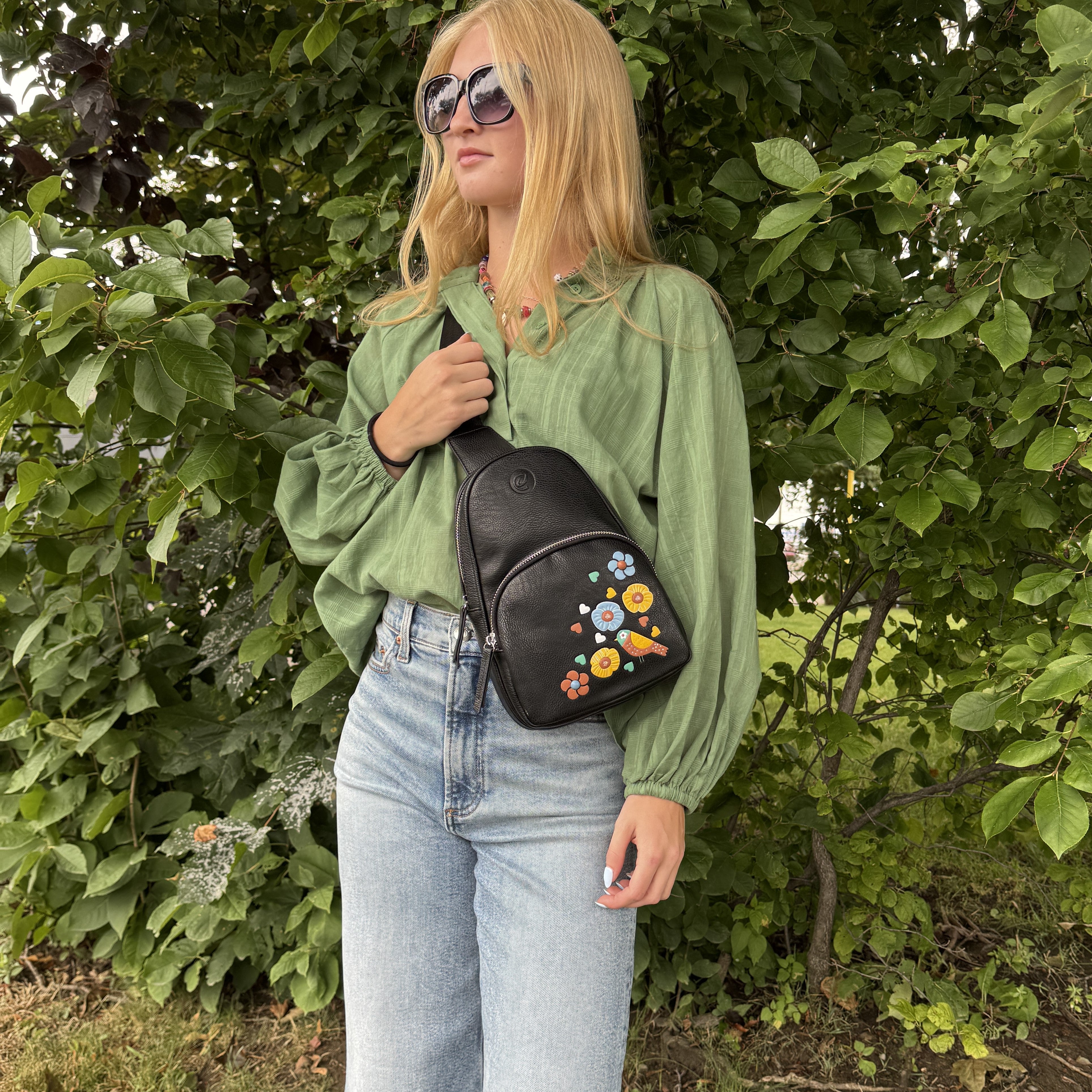 Navy Jay Sling bag floral bird detail person wearing a green shirt and blue jeans holding a black floral-patterned bag in front of green foliage.