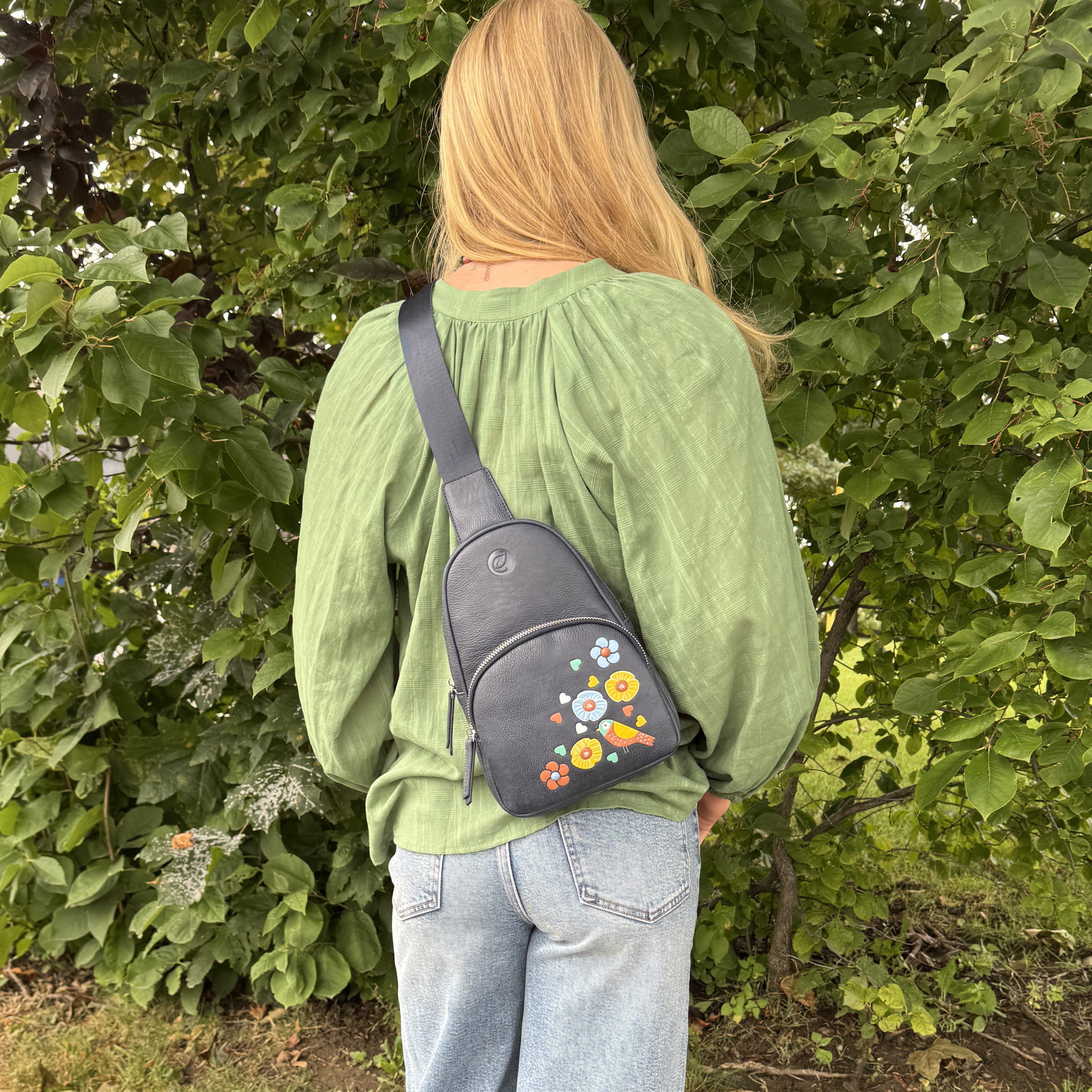Navy Jay Sling bag floral bird detail Person wearing a green shirt and light blue jeans with a gray crossbody bag featuring floral designs, standing in front of green foliage.
