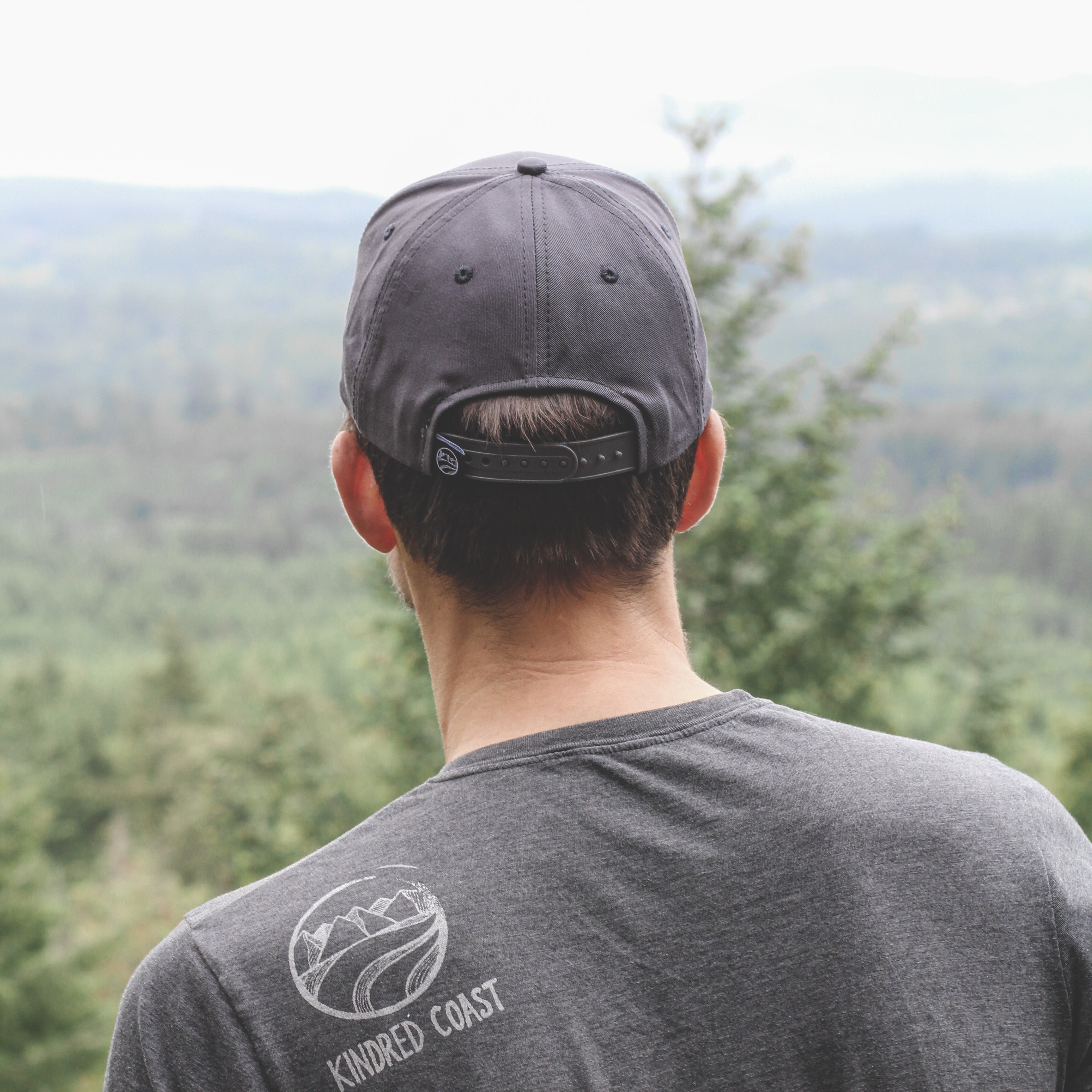 Person wearing a dark gray t-shirt with a logo and text, standing in a forested area.