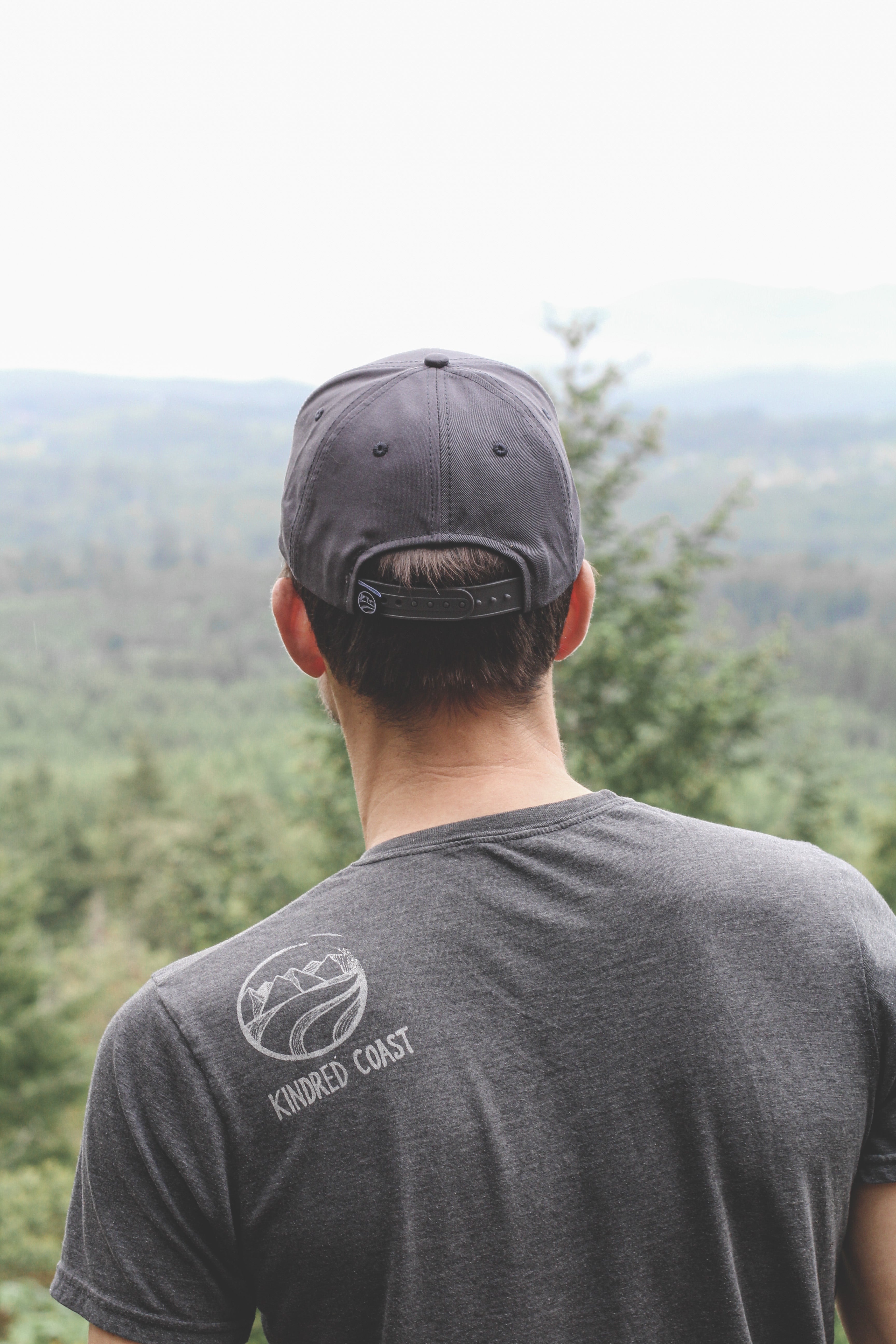 Person wearing a dark gray t-shirt with a logo and text, standing in a forested area.