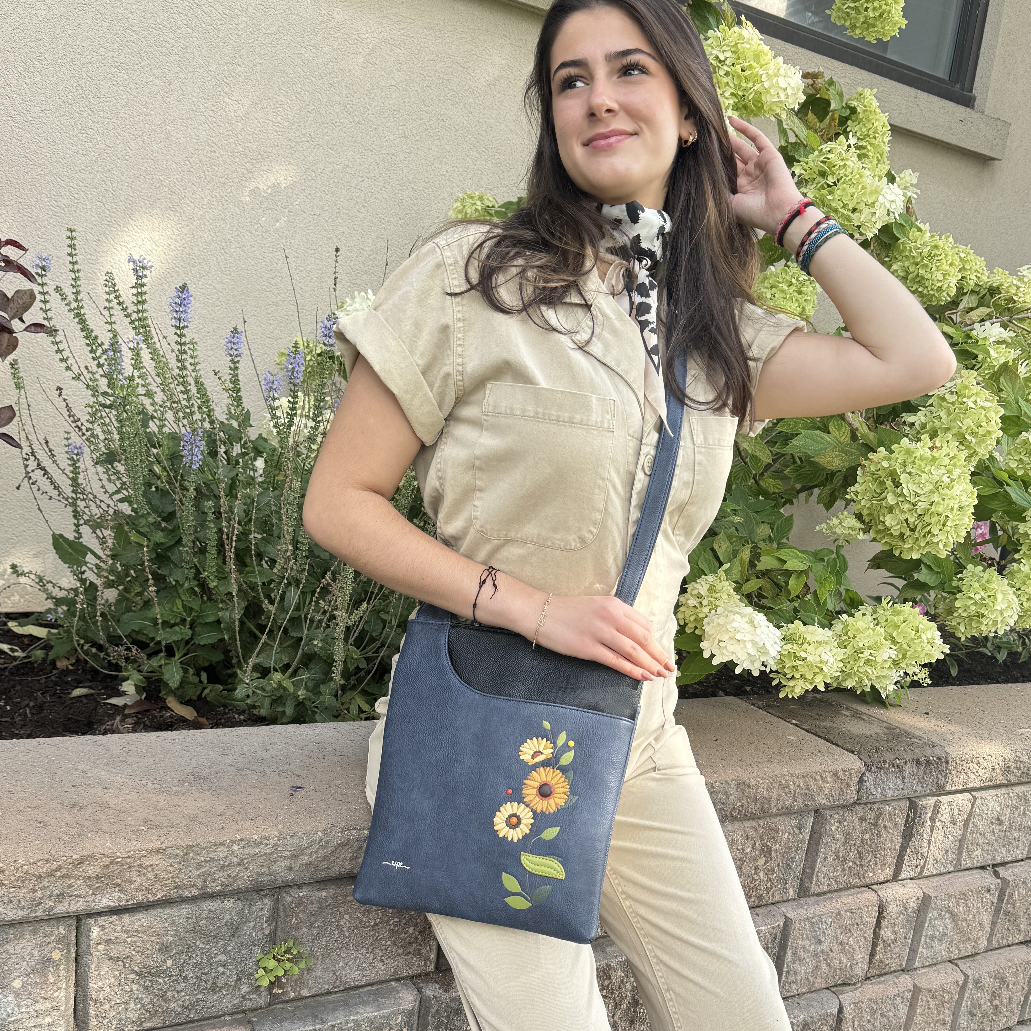 A woman wearing a beige short-sleeve shirt and matching pants carries a navy blue Espe Vegan Corp crossbody bag with a floral sunflower design on the front pocket, posed outdoors against a stone wall and green hydrangea bushes.