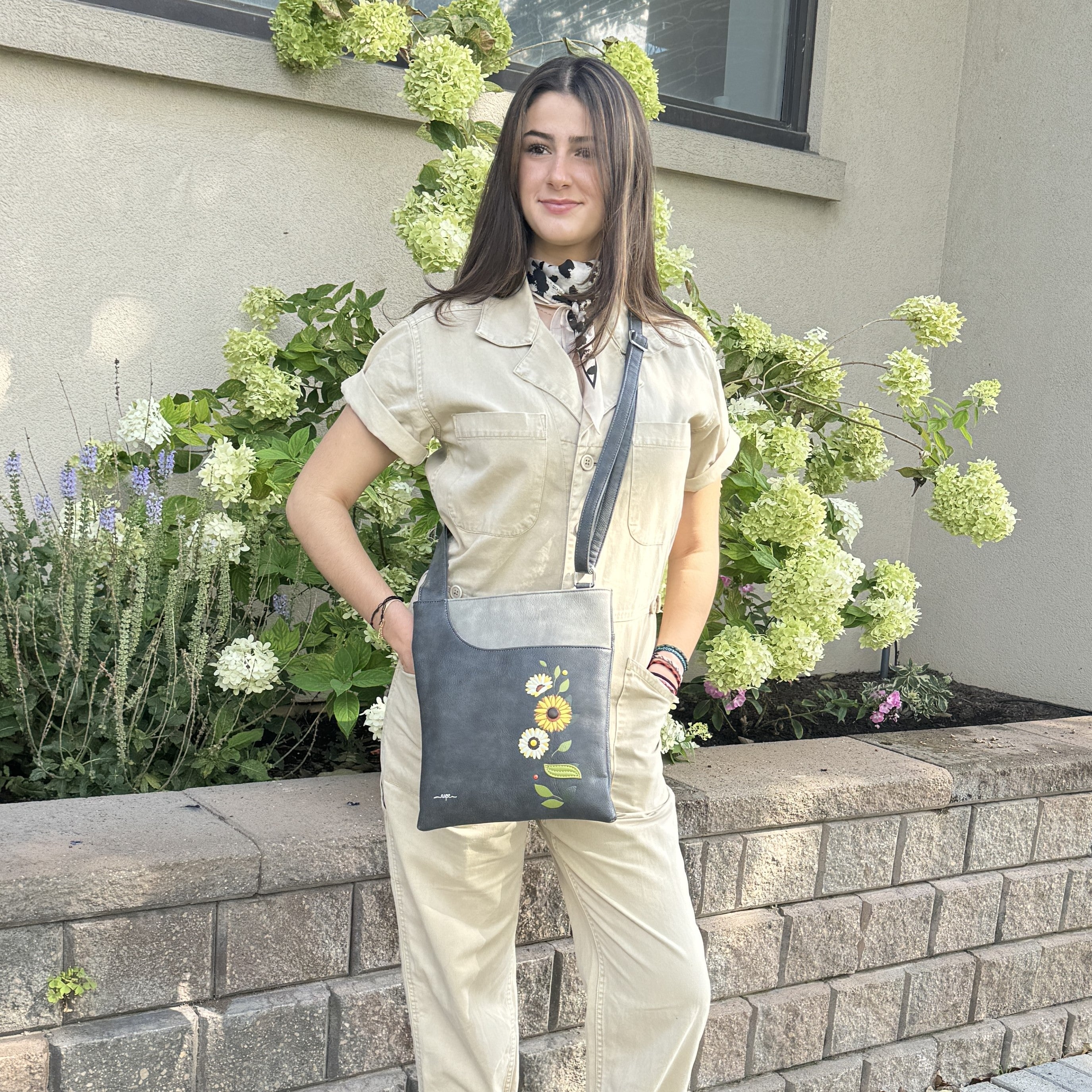 A woman wearing a beige jumpsuit stands outdoors in front of a stone wall and green hydrangea bushes, holding a dark gray Espe Vegan Corp  crossbody tote bag with a floral print.