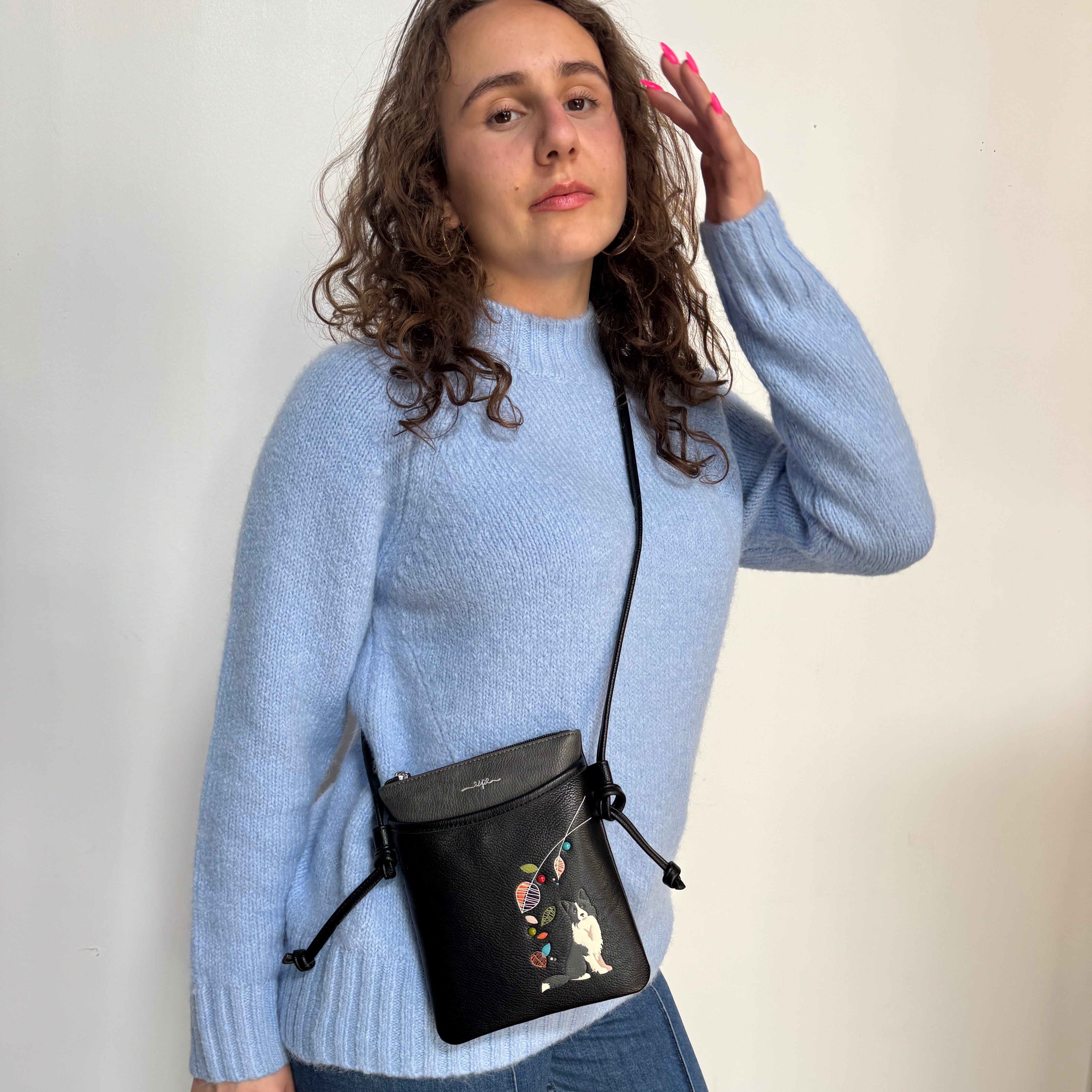 A woman wearing a light blue knit sweater and jeans models a black leather crossbody bag with a floral and cat illustration on the front pocket, set against a plain off-white wall.
