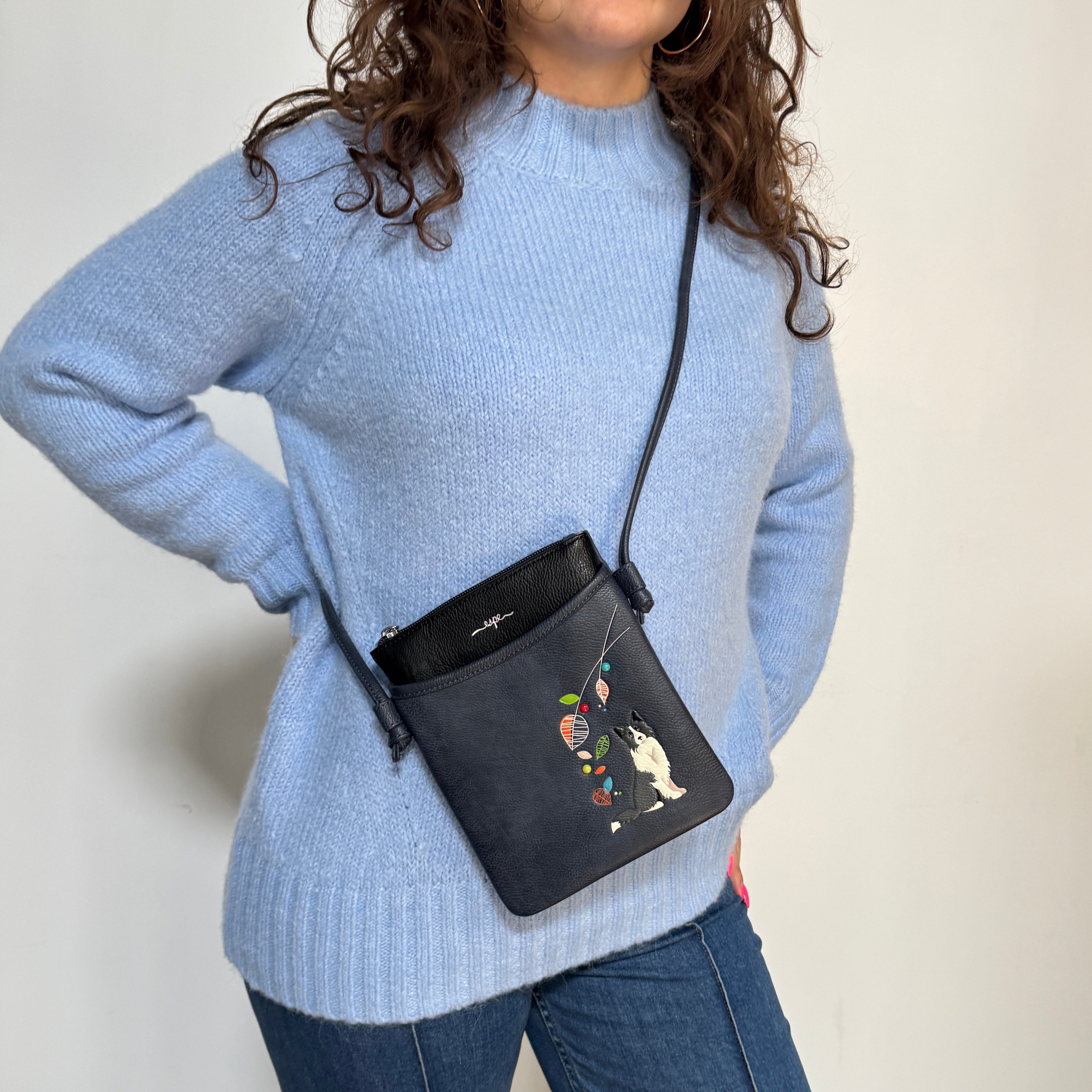 A woman wearing a light blue knit sweater and blue jeans carries a small black crossbody bag with a colorful floral and rabbit illustration on the front pocket. The photo is taken against a plain light background.