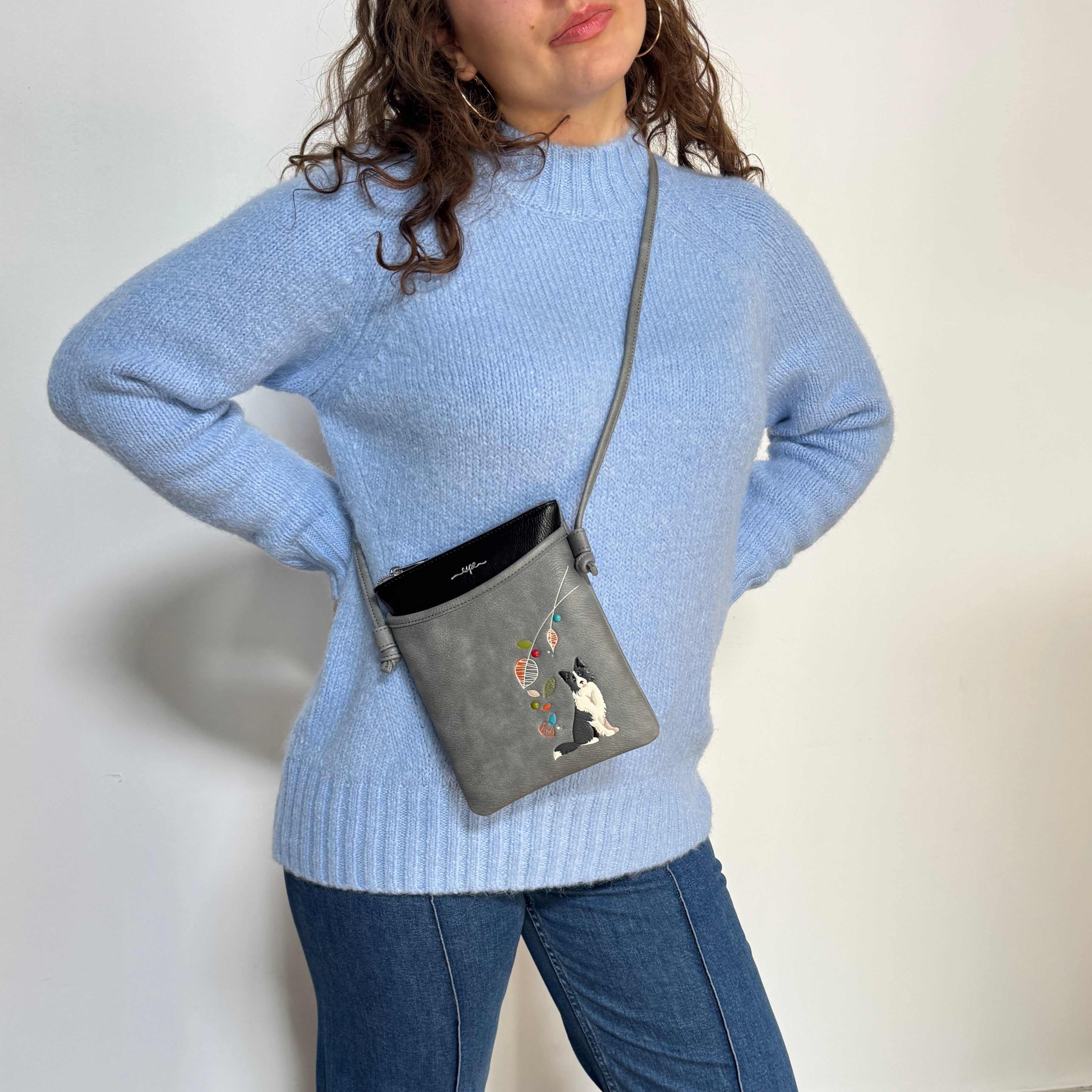 A woman wearing a light blue knit sweater and blue jeans carries a small gray crossbody bag with a cat illustration and colorful hanging balloon details. The photo is taken against a plain off-white wall.