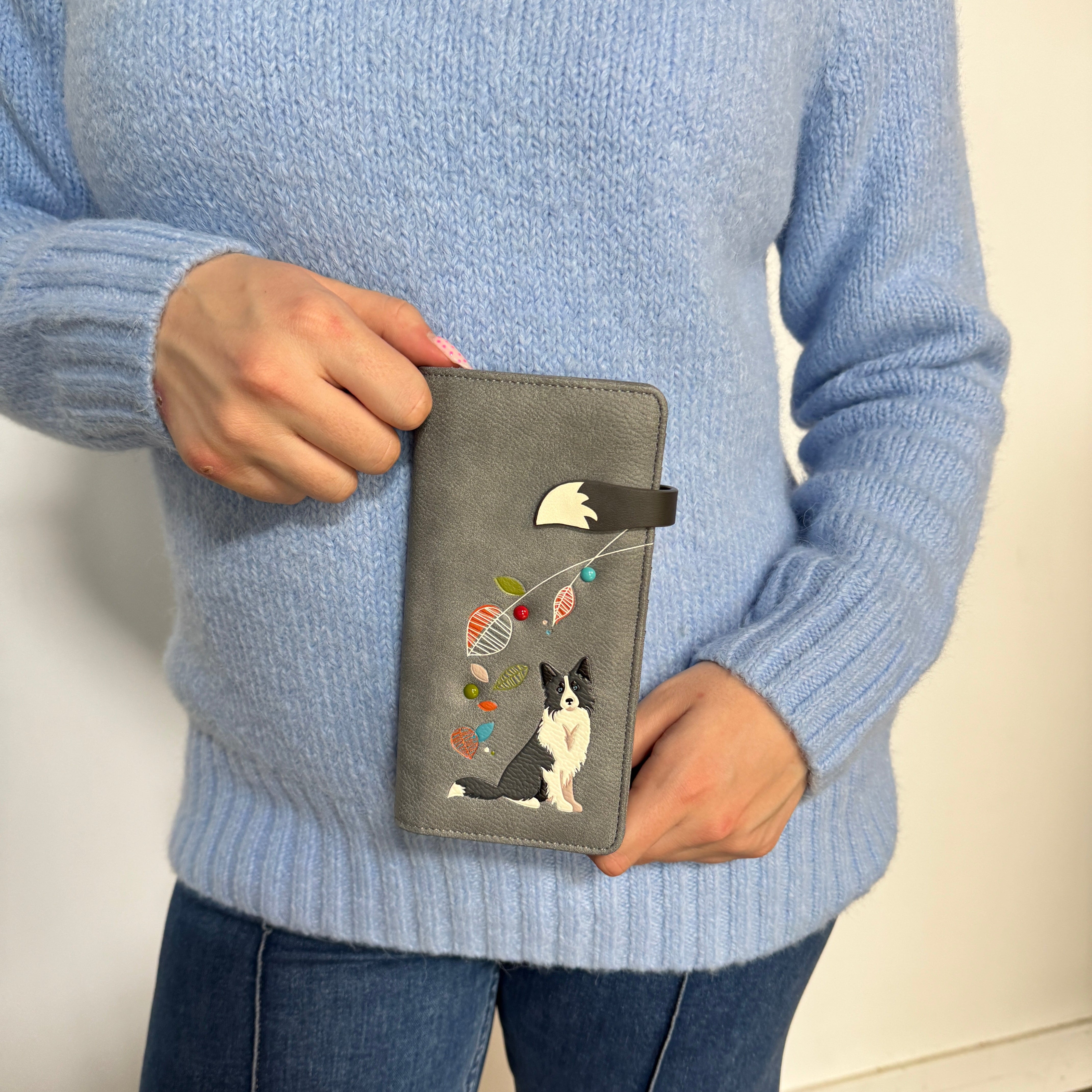 A person wearing a light blue knit sweater holds a Espe Vegan Corp grey passport cover featuring an embroidered black and white dog with colorful yarn balls.