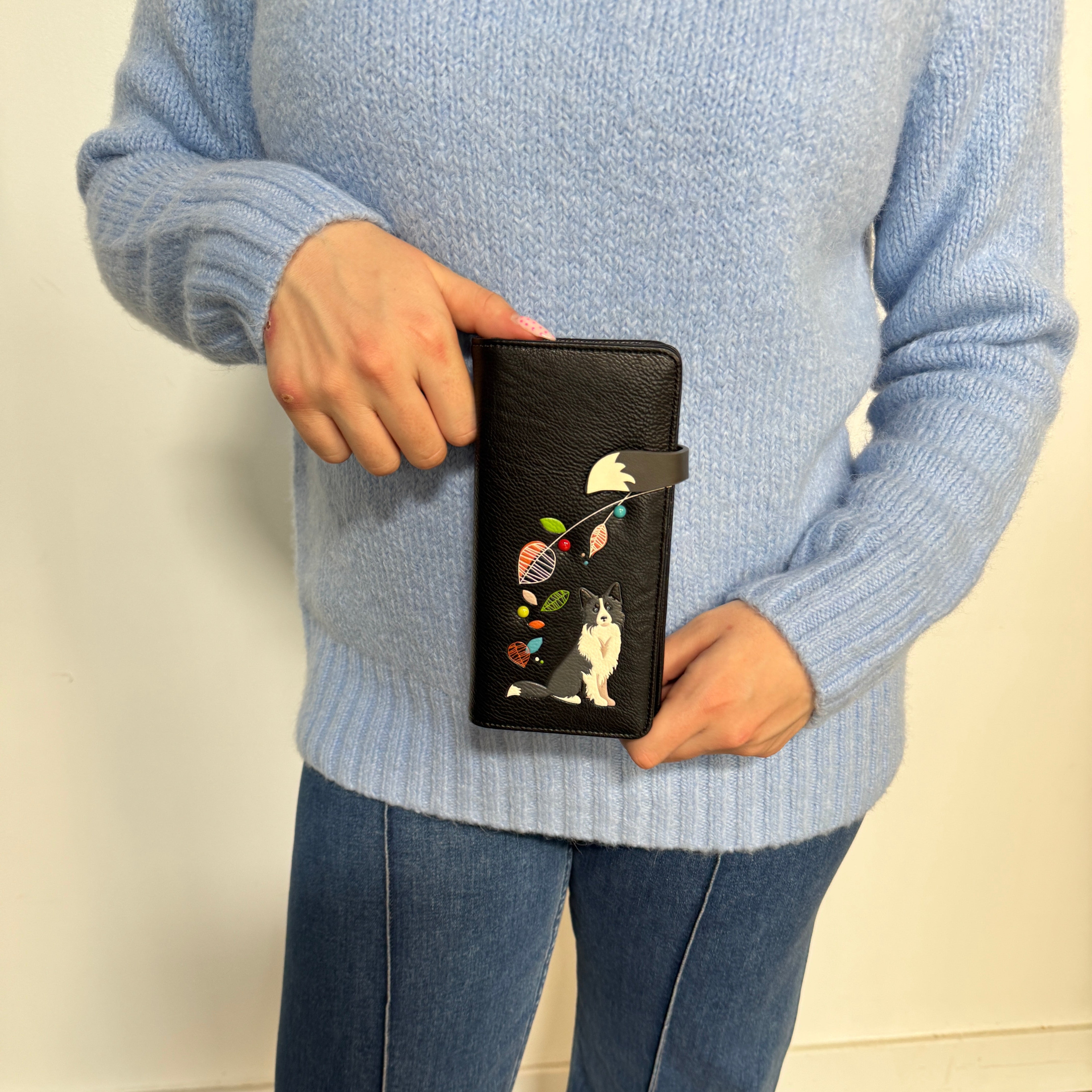 A person wearing a light blue ribbed knit sweater and blue jeans holds a Espe Vegan Corp black textured wallet or clutch with a colorful embroidered cat design and small floral details.