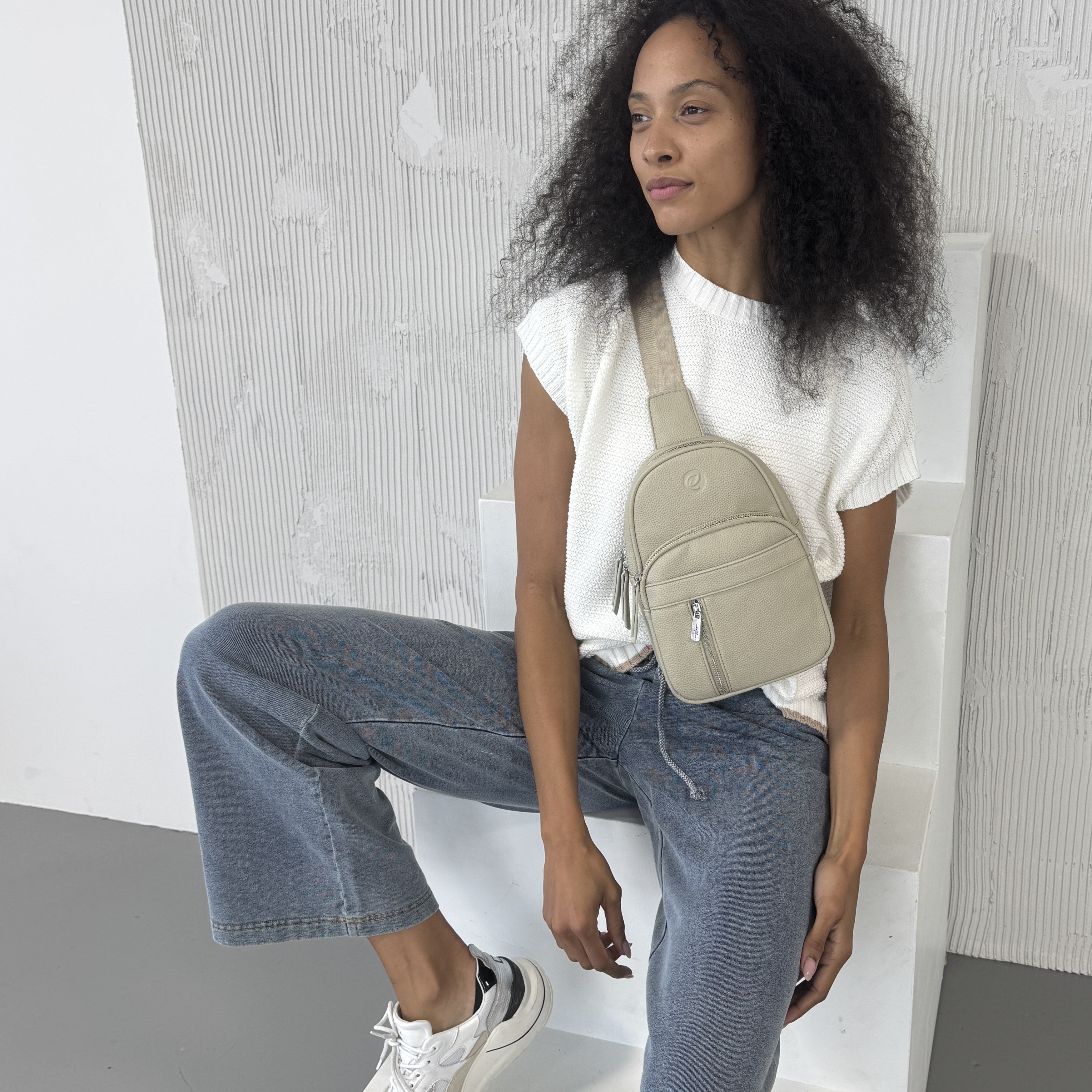 Woman sitting on a white chair wearing a white top, blue jeans, and a Espe Vegan Corp beige crossbody bag.