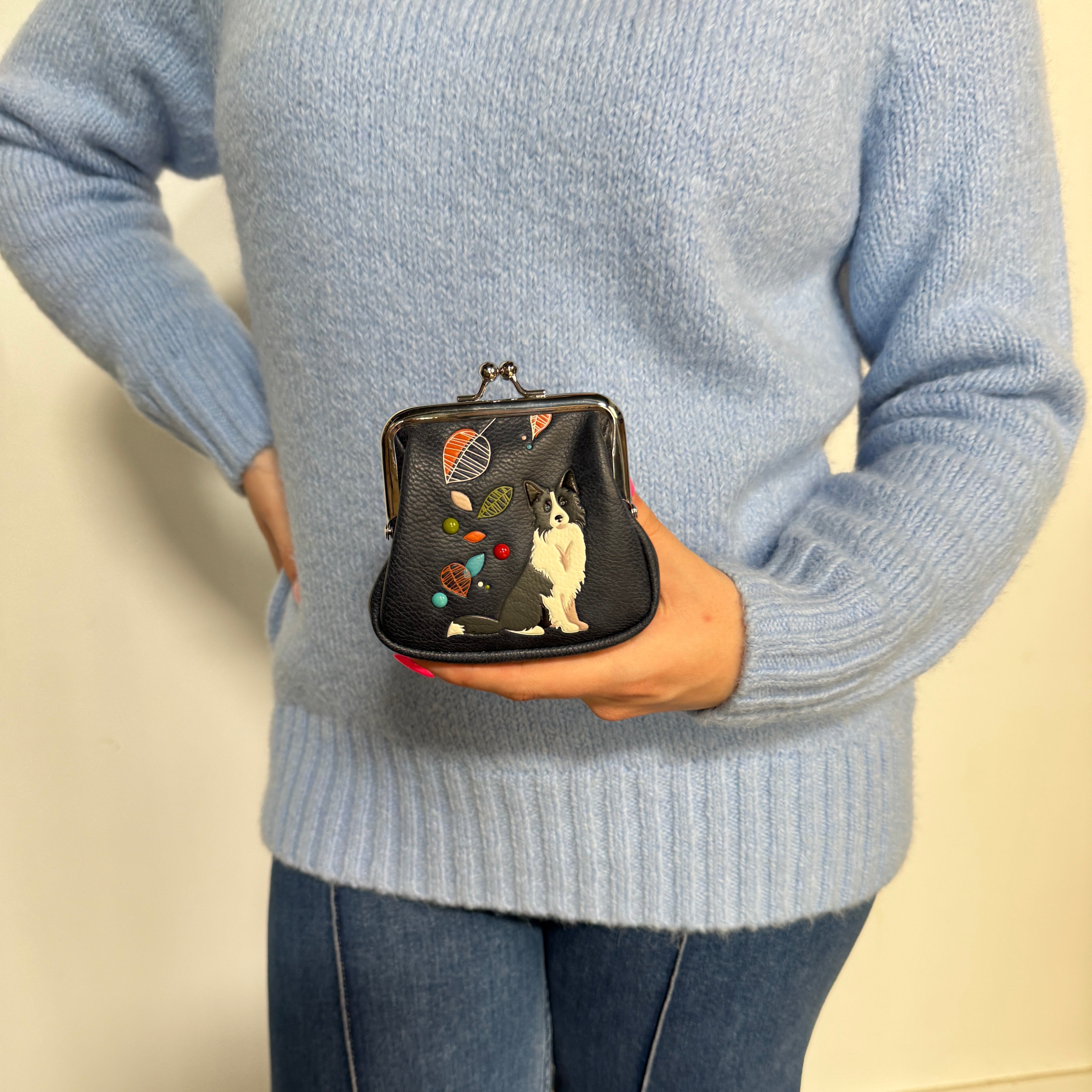 Espe Vegan Corp small black kiss-lock coin purse decorated with a black-and-white dog and colorful abstract shapes, paired with blue jeans against a plain light beige wall.
