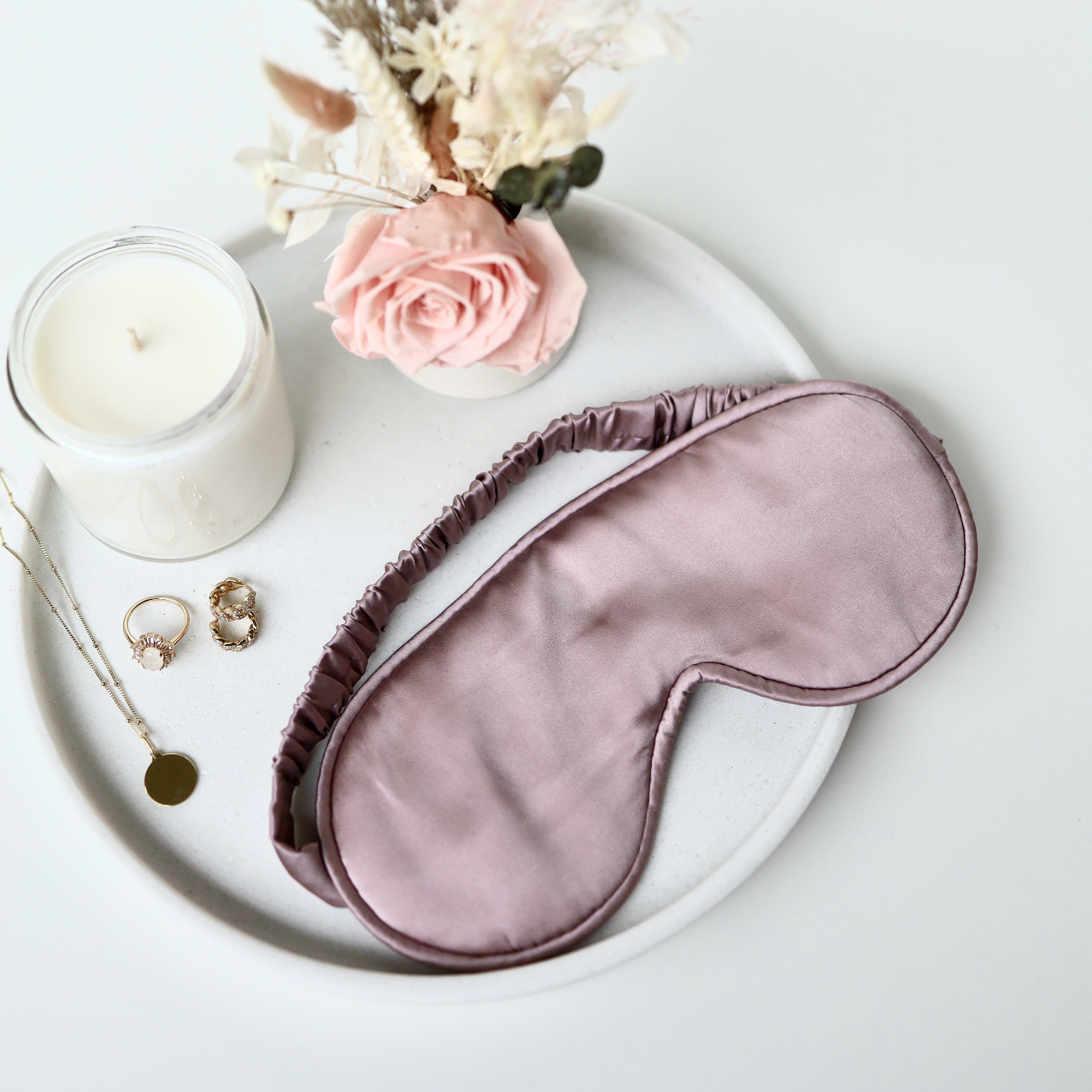 Pink satin eye mask on a white plate with a candle, flowers, and jewelry.