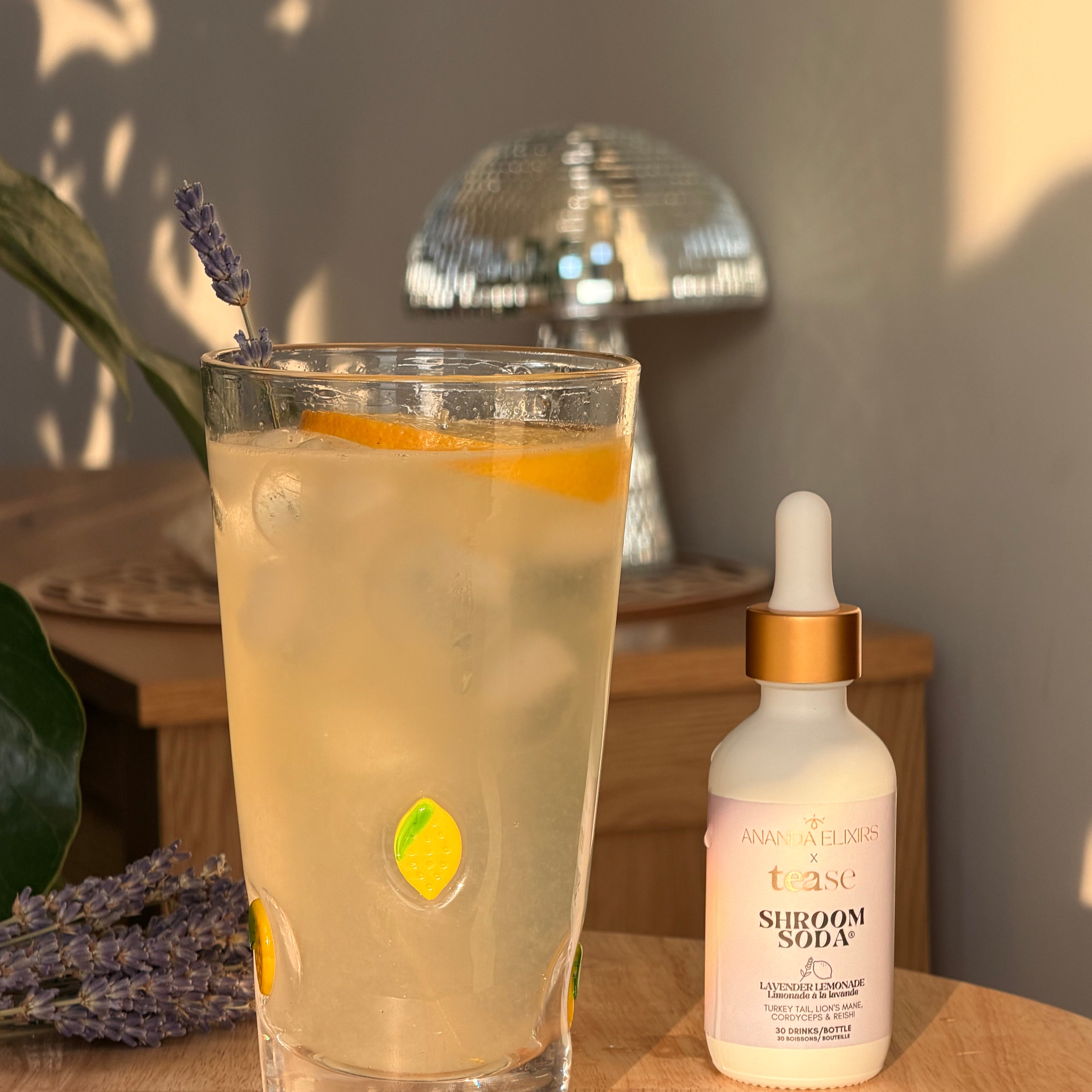 Ananda Elixirs Shroom Soda® – Lavender Lemonade – Sparkling mocktail with lemon slice, lavender sprig, and dropper bottle on wooden table