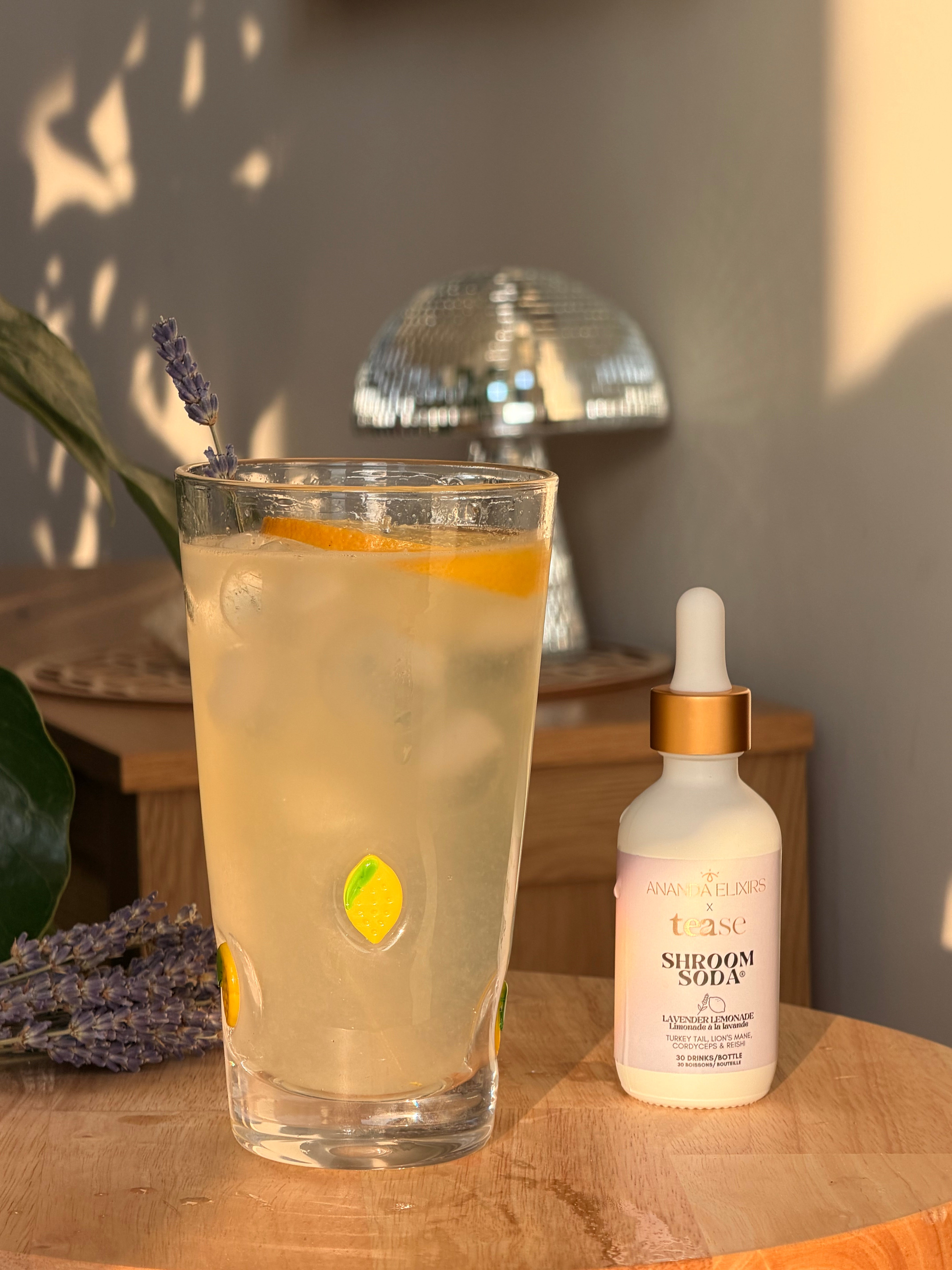 Ananda Elixirs Shroom Soda® – Lavender Lemonade – Sparkling mocktail with lemon slice, lavender sprig, and dropper bottle on wooden table