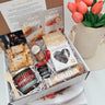 Gift box with Cama products including cheese, jam, and other snacks, accompanied by a vase of tulips.