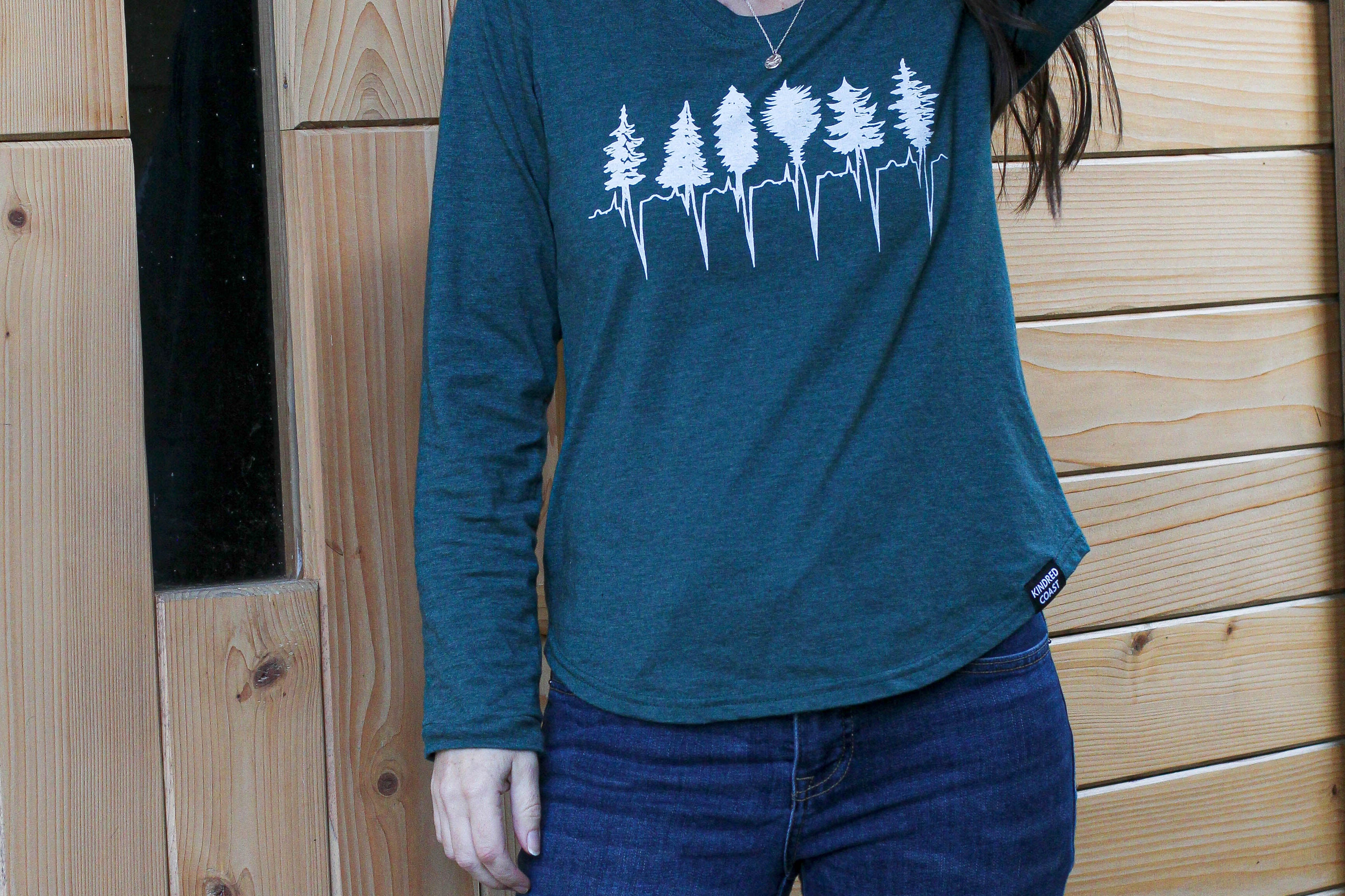 Woman wearing a teal long-sleeve shirt with a forest design and blue jeans, standing against a wooden wall.