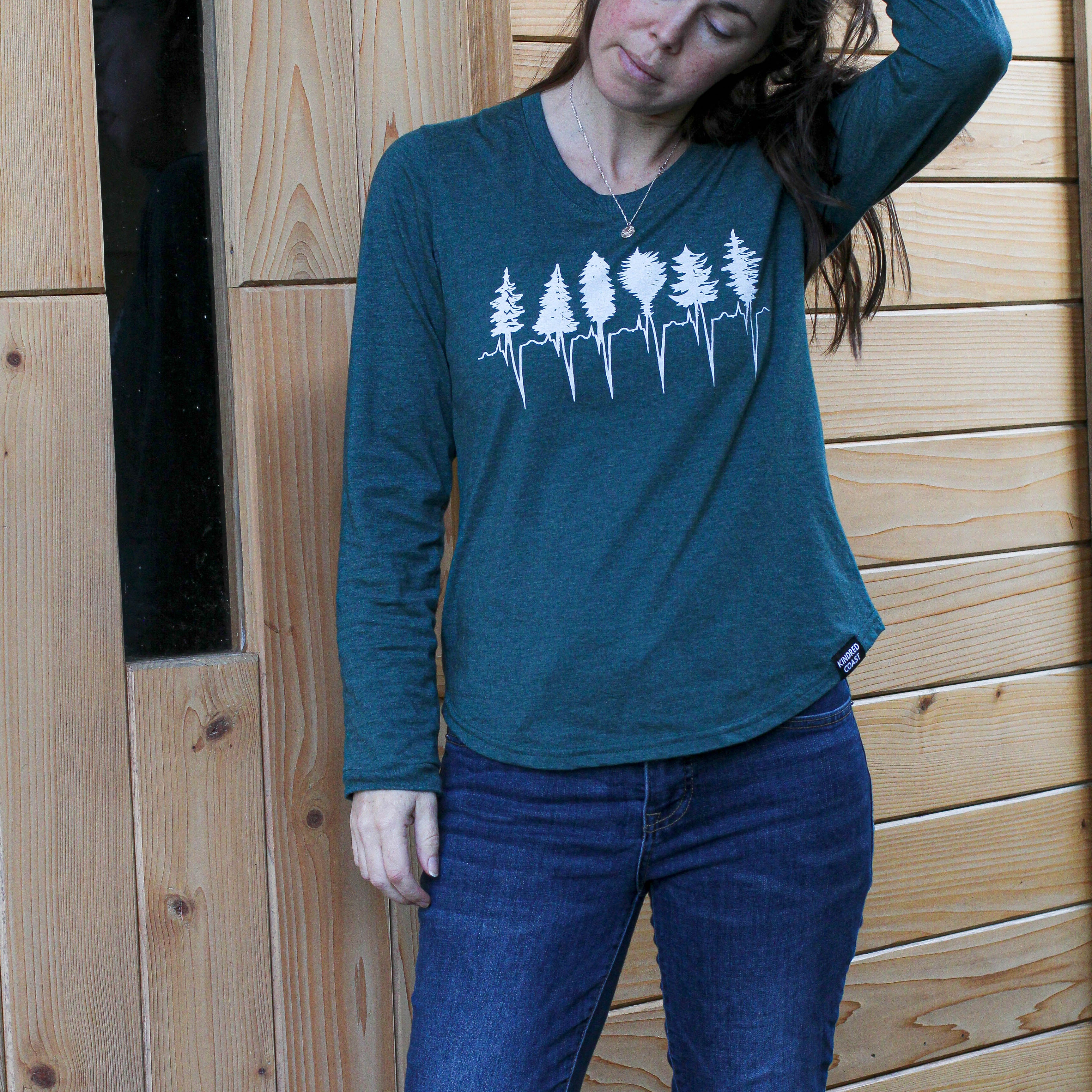 Woman wearing a teal long-sleeve shirt with a forest design and blue jeans, standing against a wooden wall.