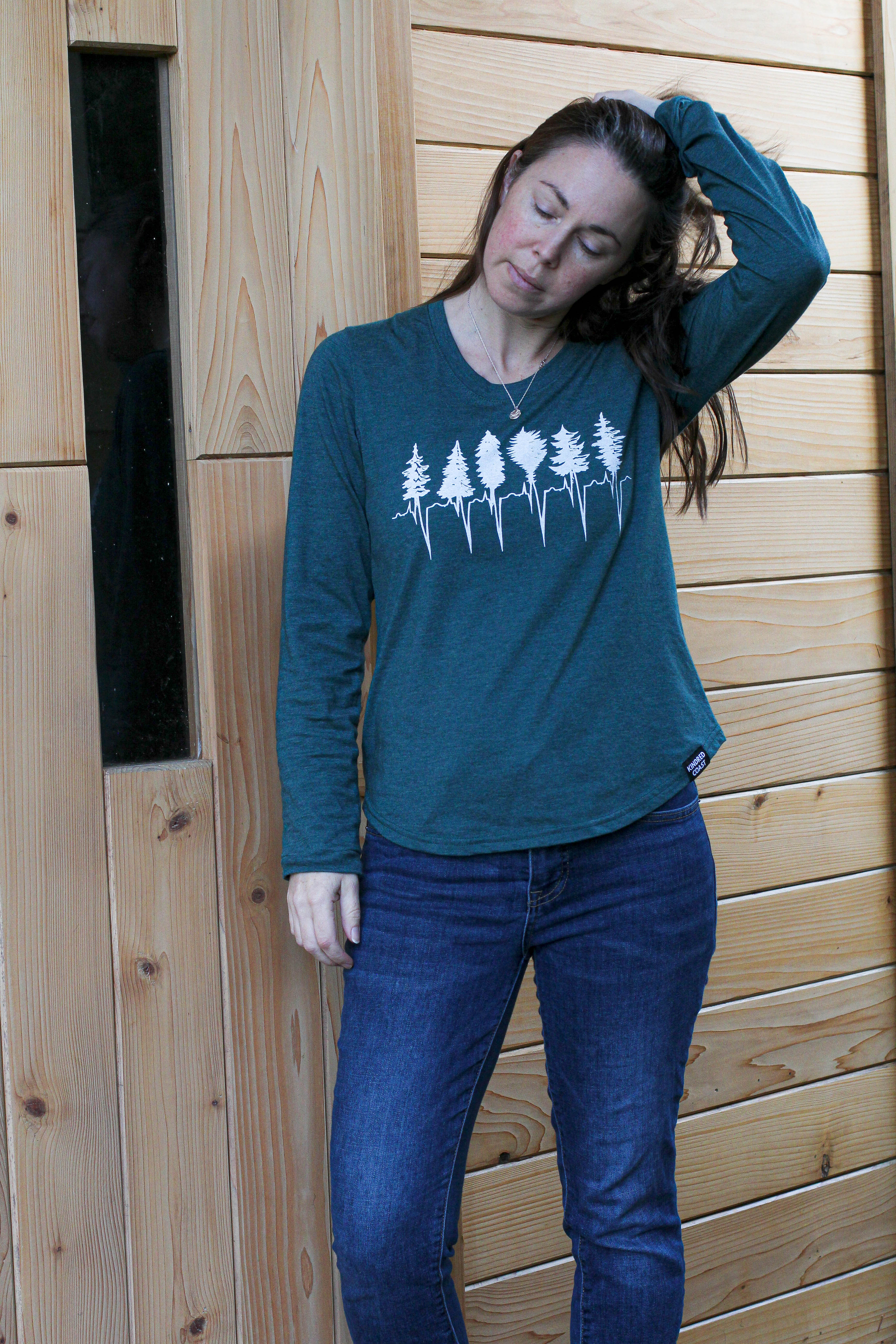 Woman wearing a teal long-sleeve shirt with a forest design and blue jeans, standing against a wooden wall.
