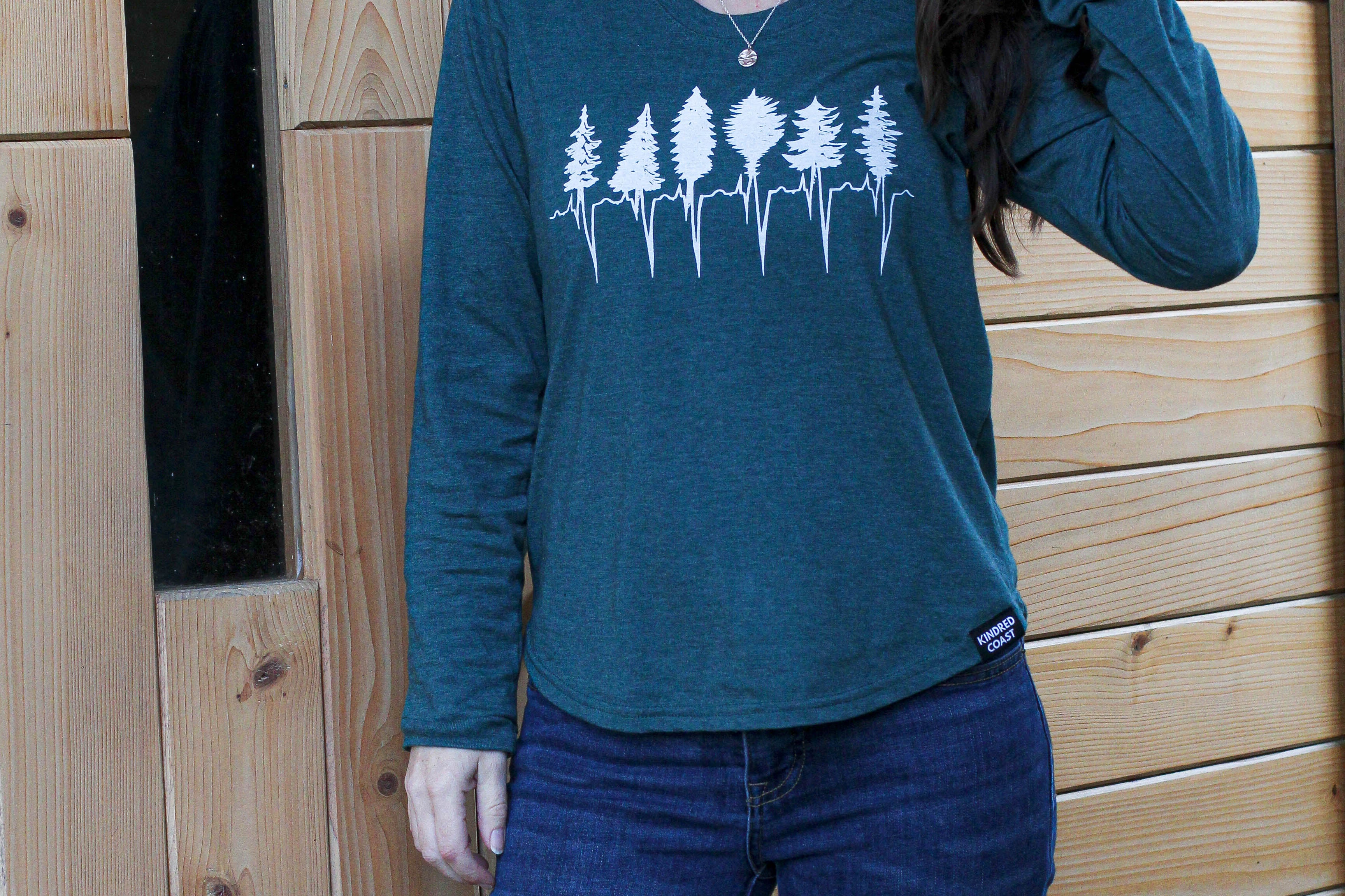 Woman wearing a teal long-sleeve shirt with a forest design against a wooden wall.