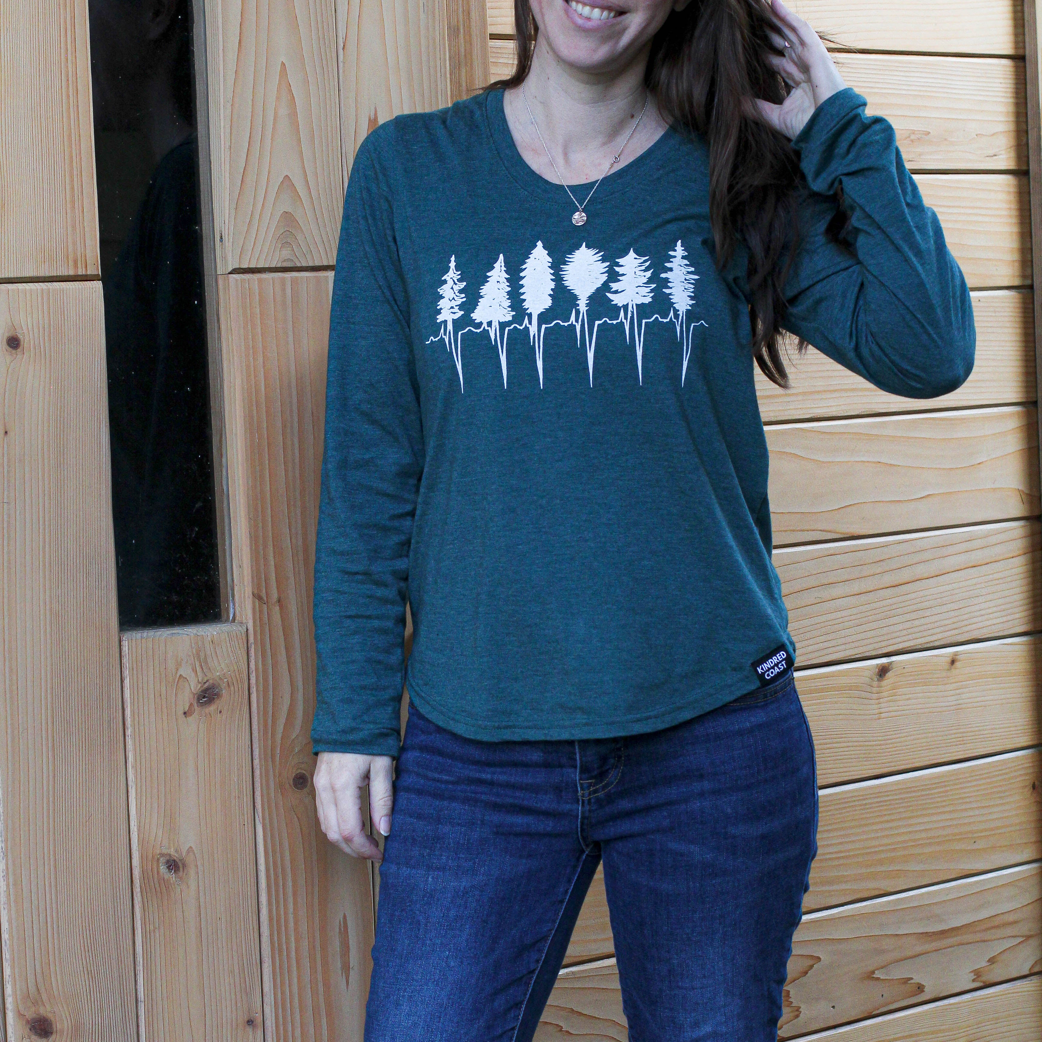 Woman wearing a teal long-sleeve shirt with a forest design against a wooden wall.