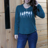 Woman wearing a teal long-sleeve shirt with a forest design against a wooden wall.