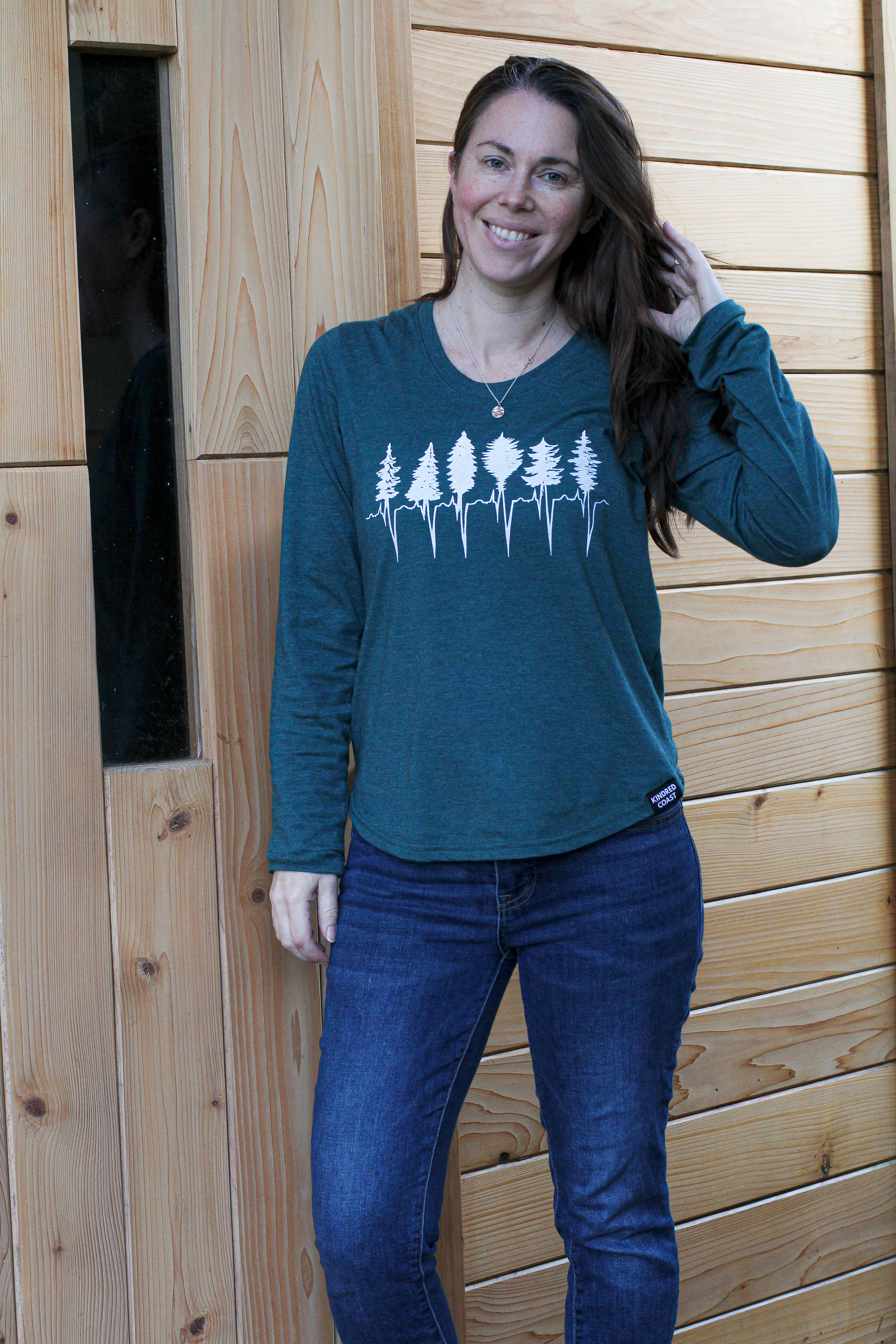 Woman wearing a teal long-sleeve shirt with a forest design against a wooden wall.