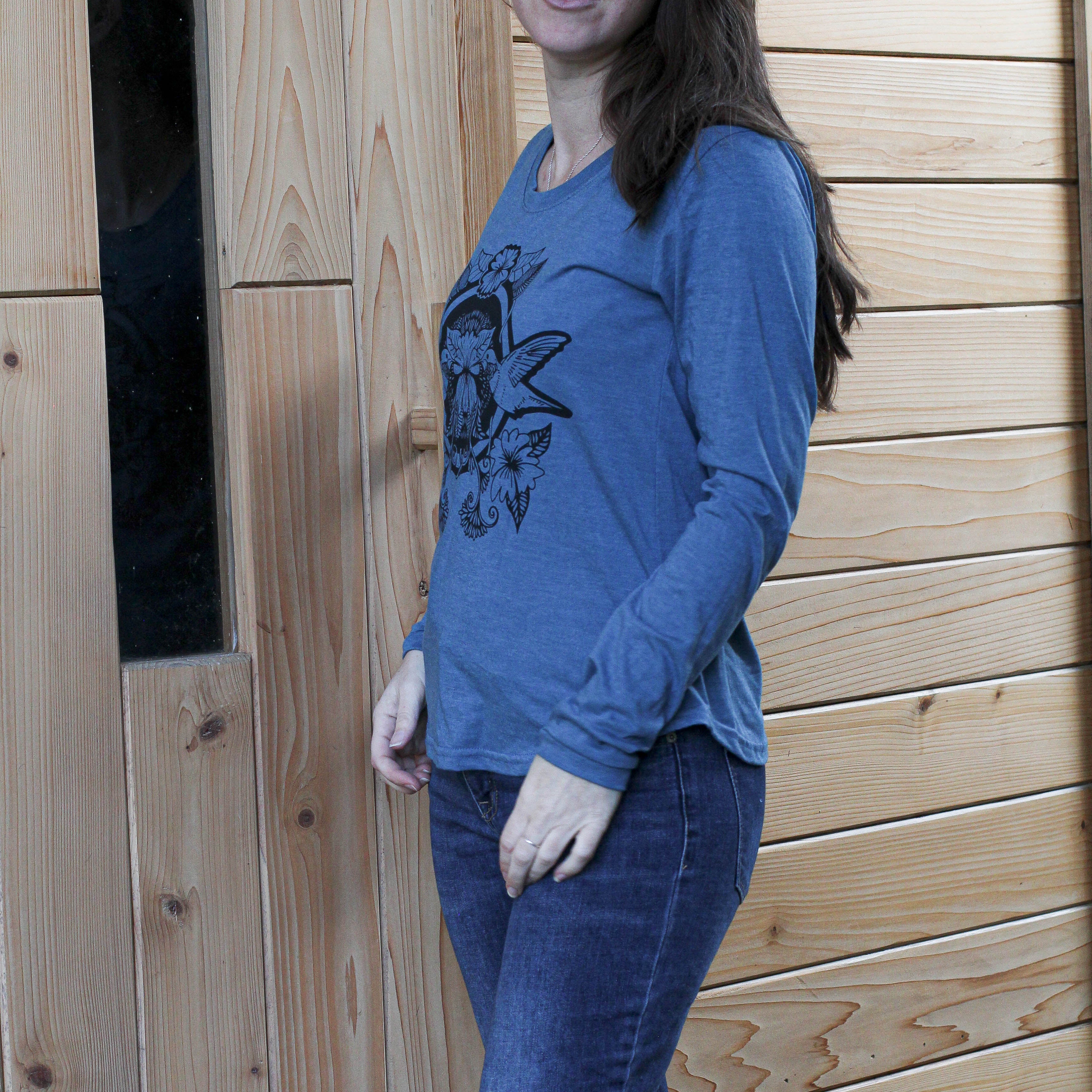 Woman wearing a blue sweater and jeans standing against a wooden wall.