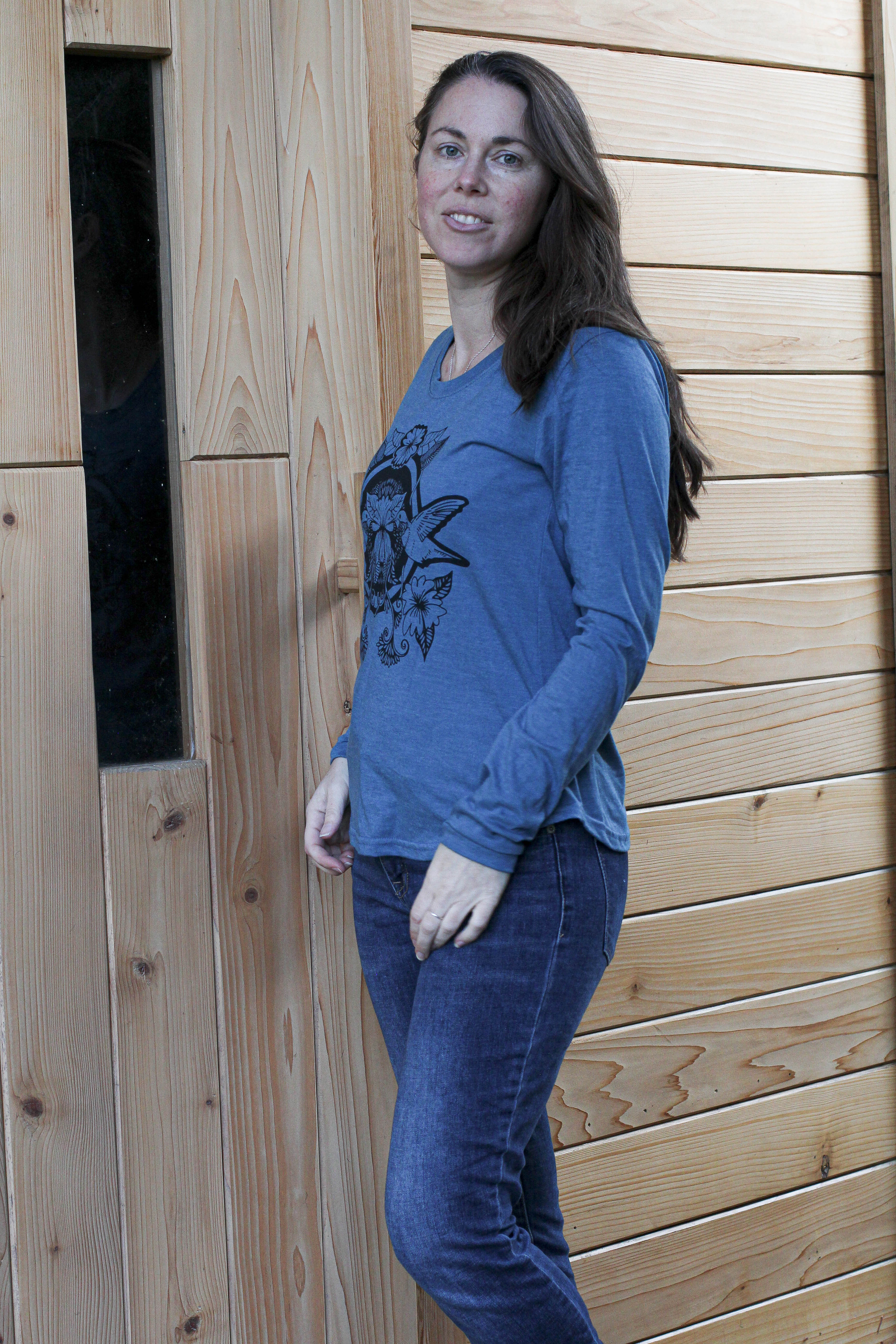 Woman wearing a blue sweater and jeans standing against a wooden wall.