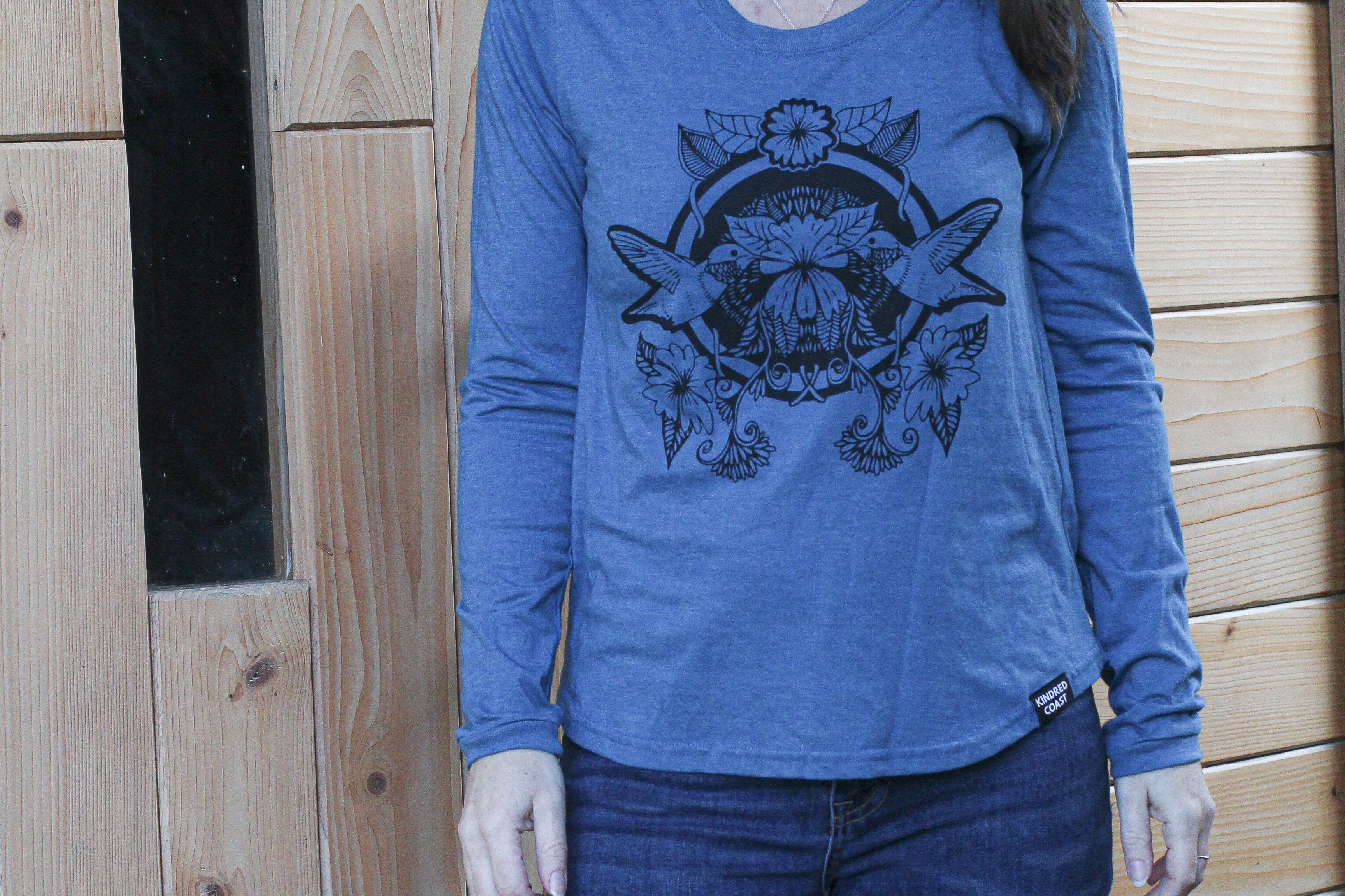 Woman wearing a blue long-sleeve shirt with a graphic design in front of a wooden wall.