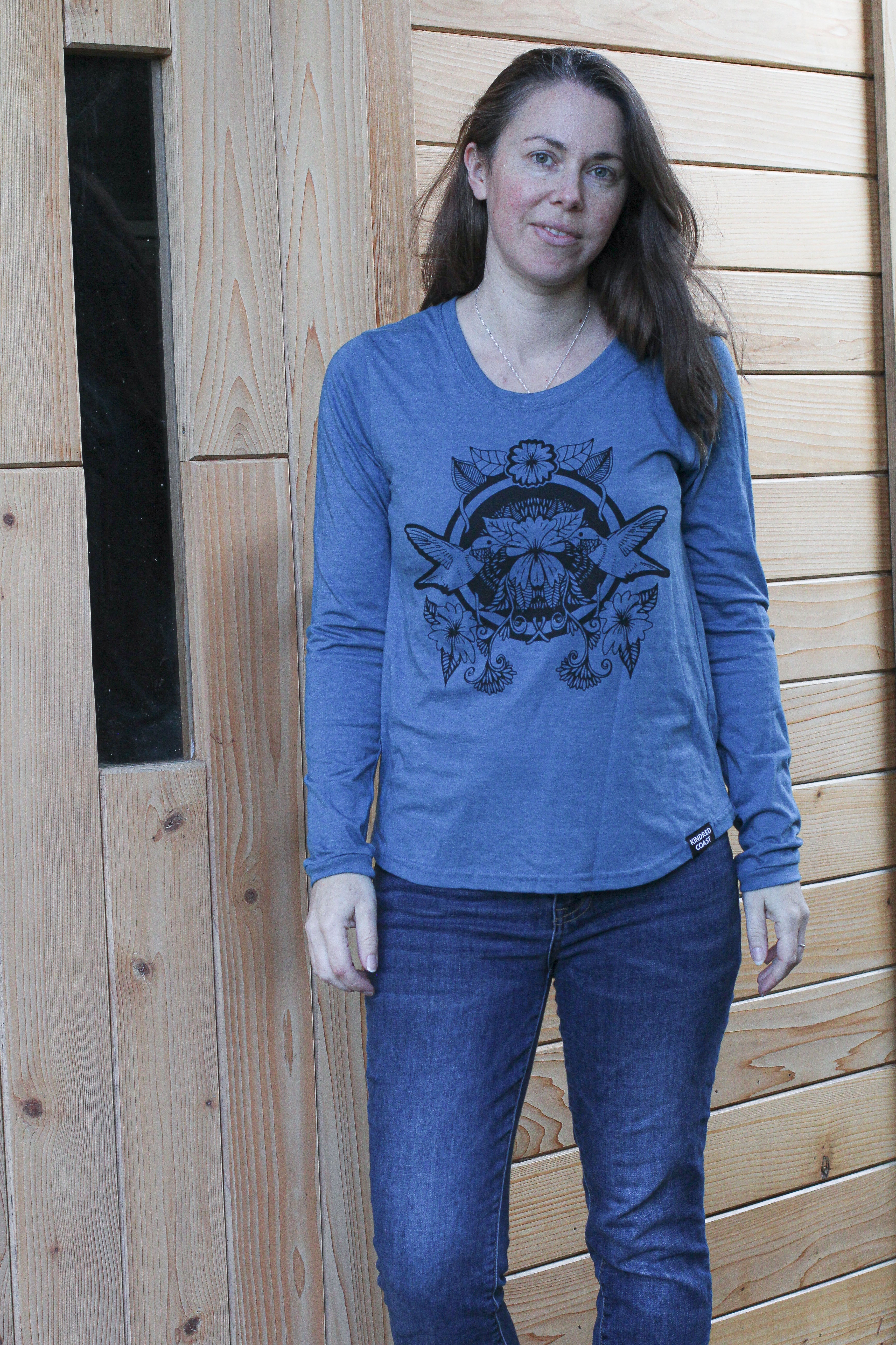 Woman wearing a blue long-sleeve shirt with a graphic design in front of a wooden wall.