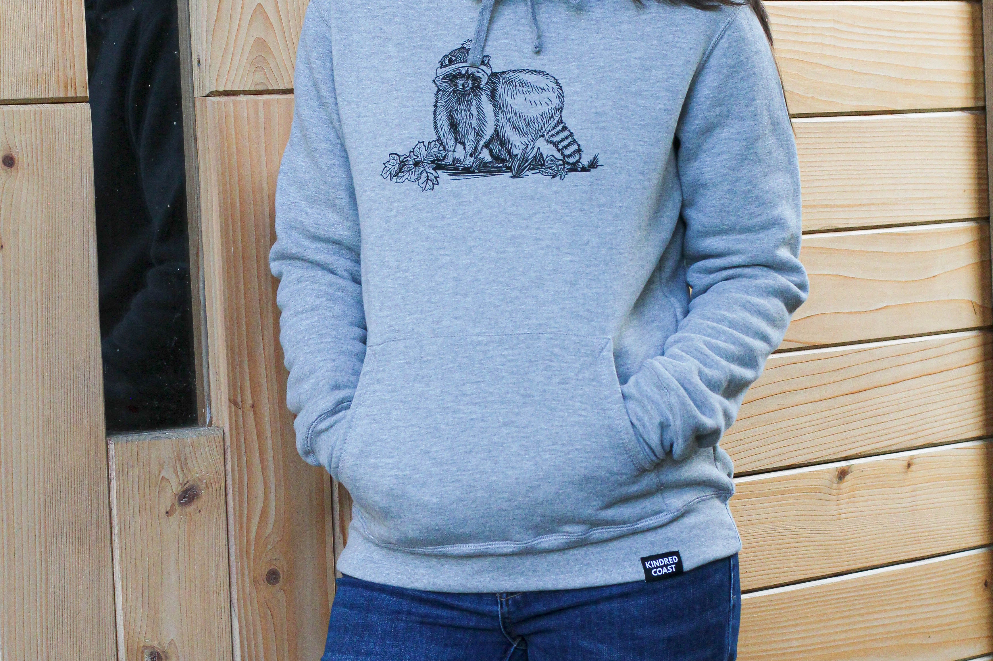 Woman wearing a gray hoodie with a graphic design standing in front of a wooden wall.