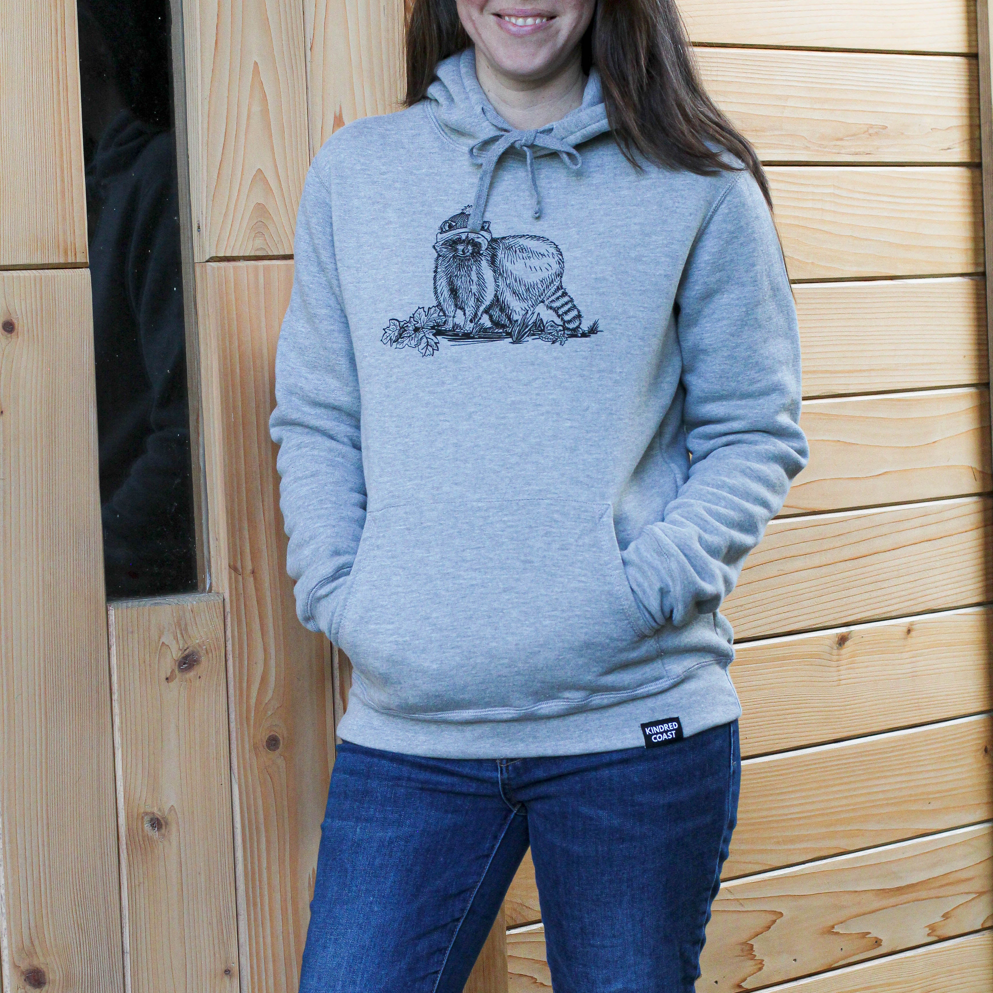 Woman wearing a gray hoodie with a graphic design standing in front of a wooden wall.