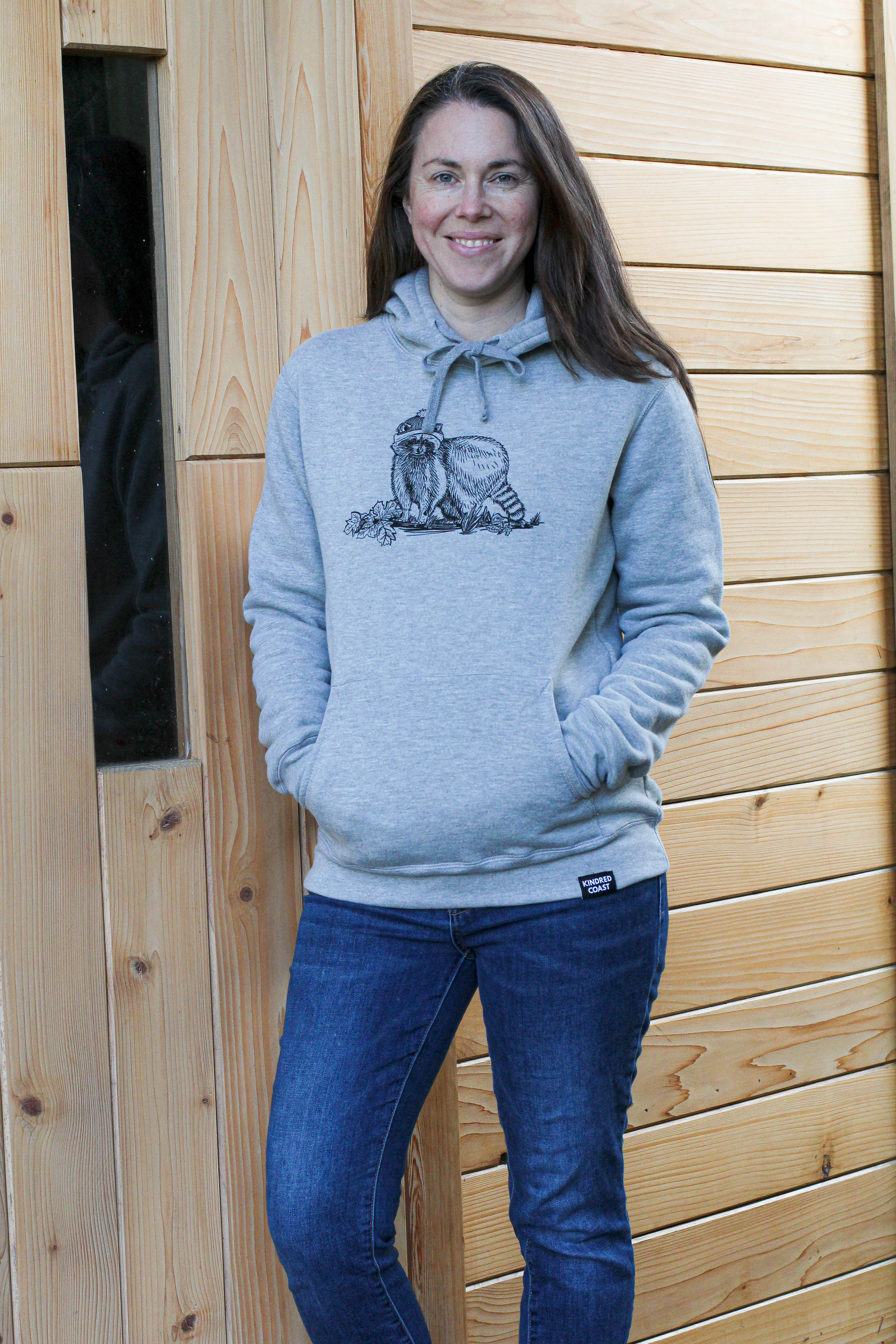 Woman wearing a gray hoodie with a graphic design standing in front of a wooden wall.