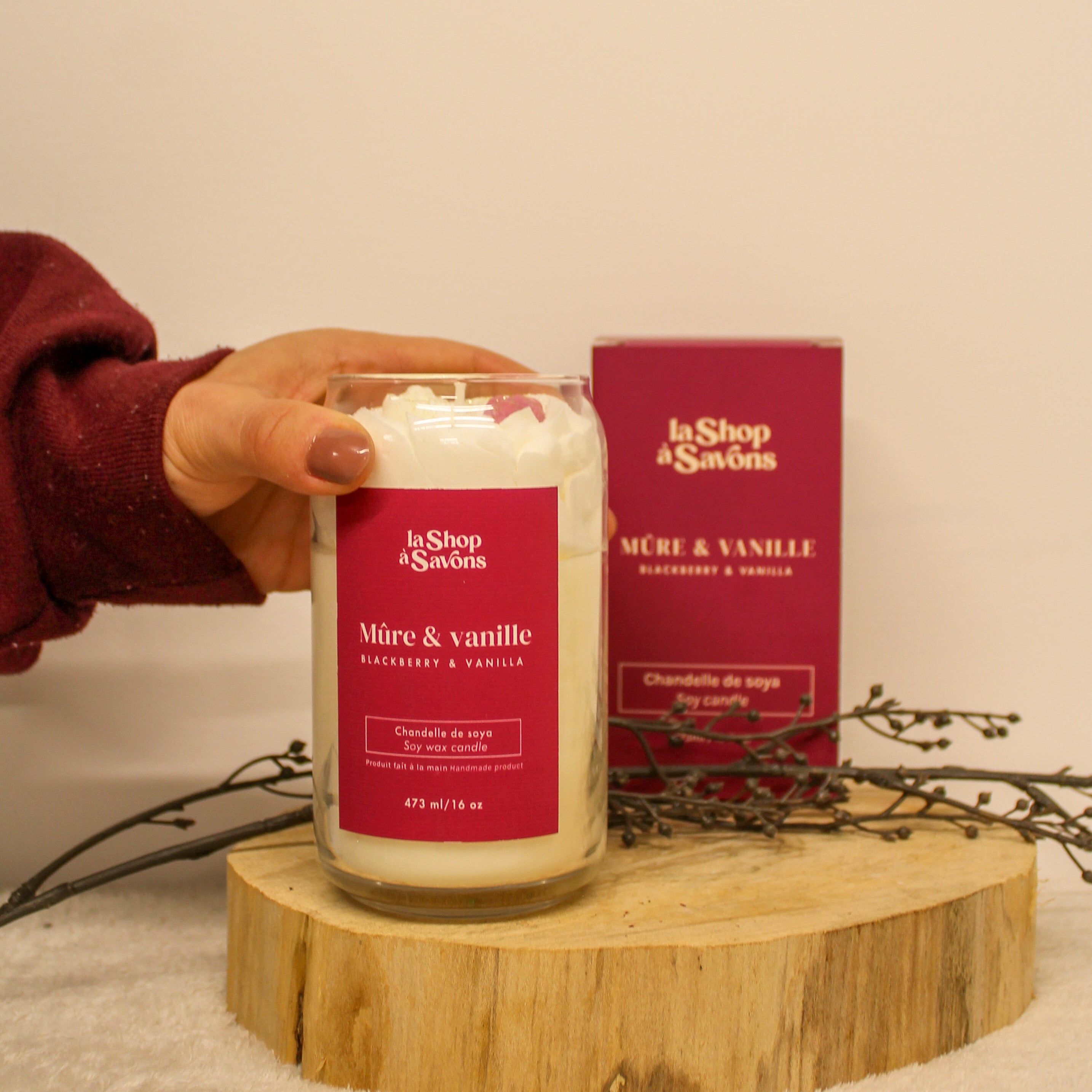 Candle with red label held by a hand on a wooden stand, with 'la Shop dé Savons' packaging in the background.