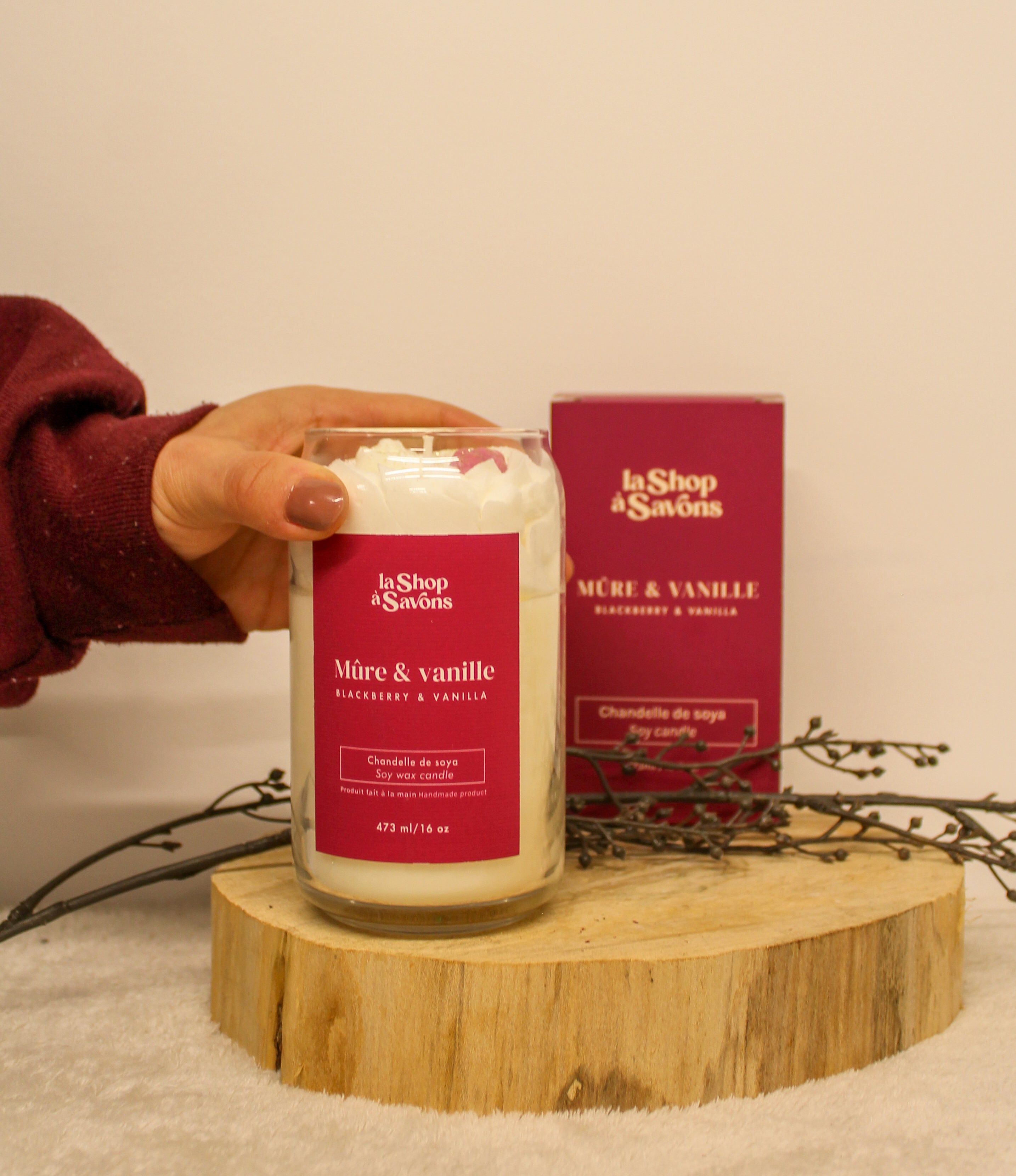 Candle with red label held by a hand on a wooden stand, with 'la Shop dé Savons' packaging in the background.