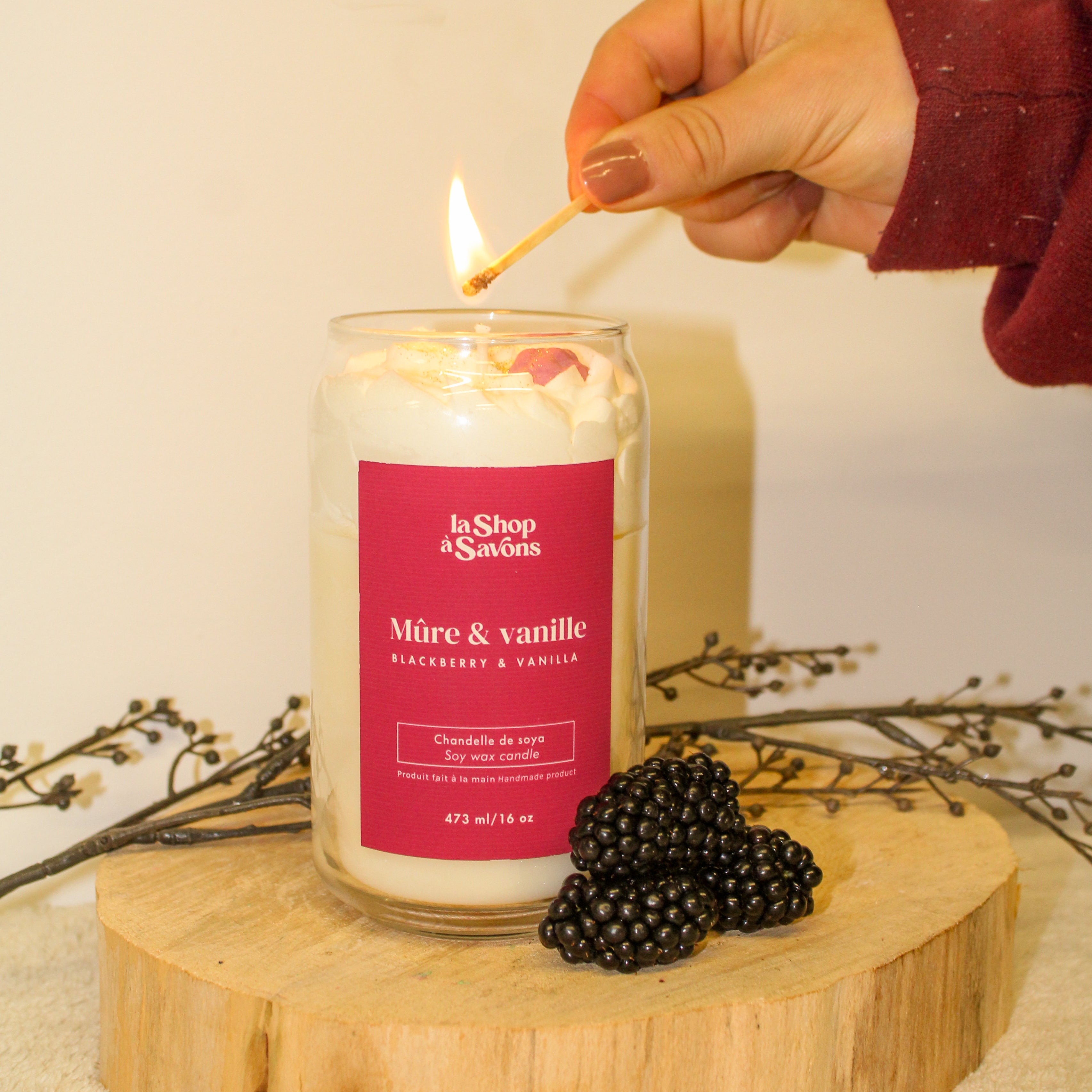Person lighting a candle labeled 'La Shop de Sables' with blackberries on a wooden stand.