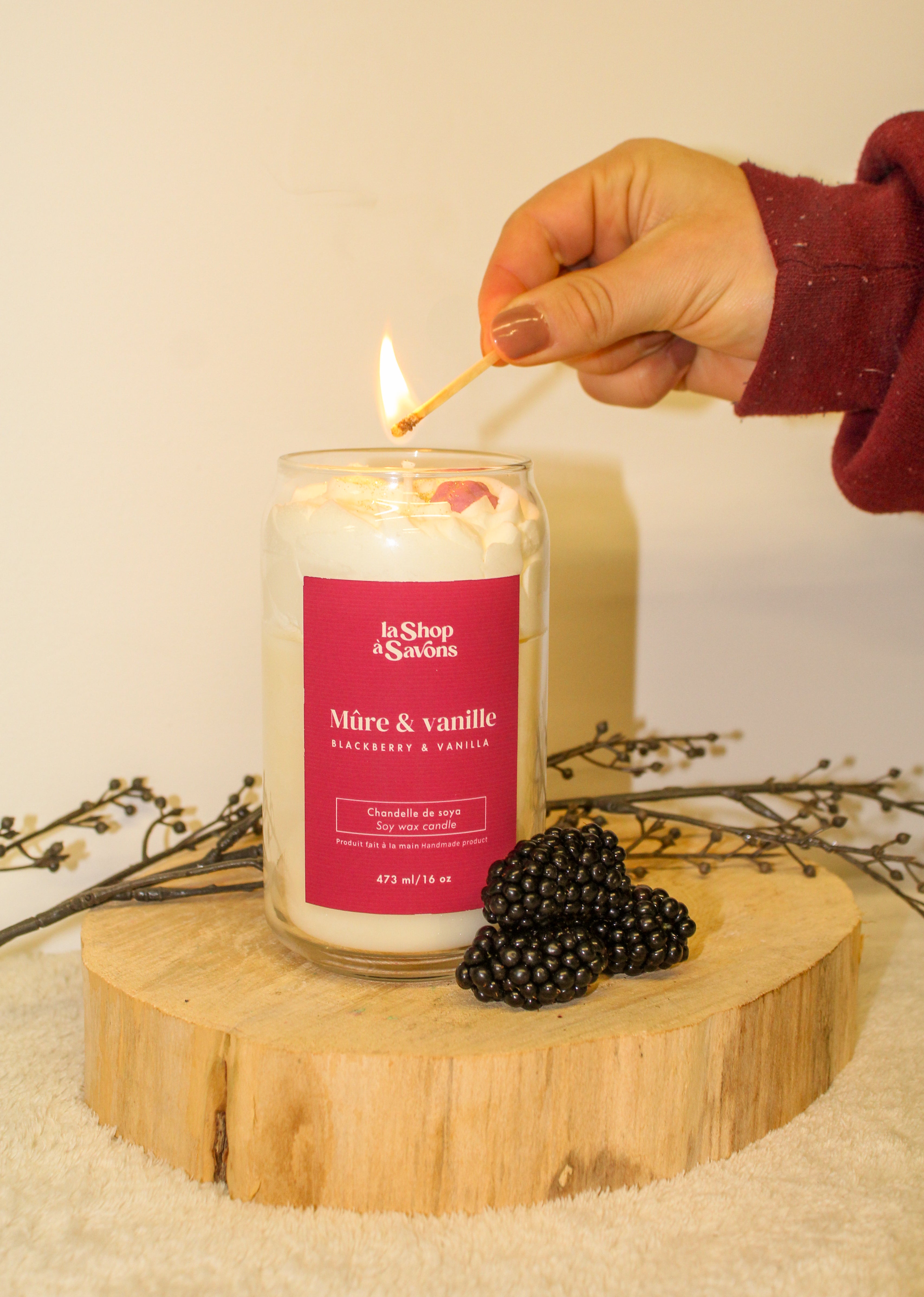 Person lighting a candle labeled 'La Shop de Sables' with blackberries on a wooden stand.