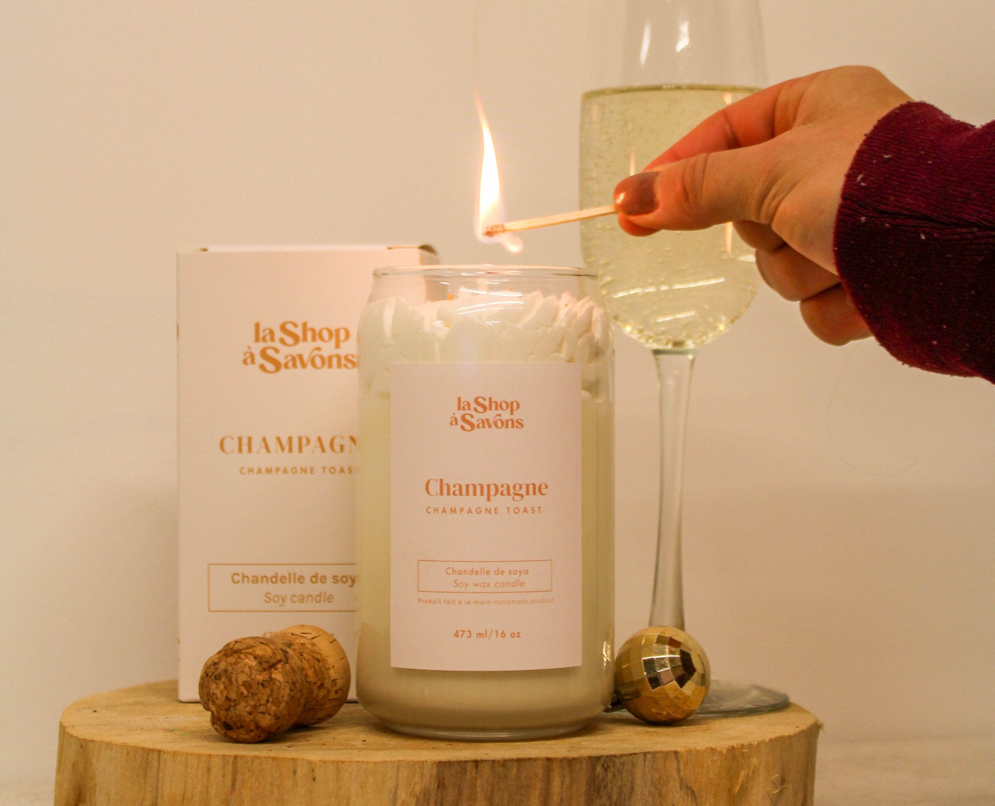 Candle labeled 'Champagne' being lit with a match, next to a glass of champagne and two corks on a wooden block.