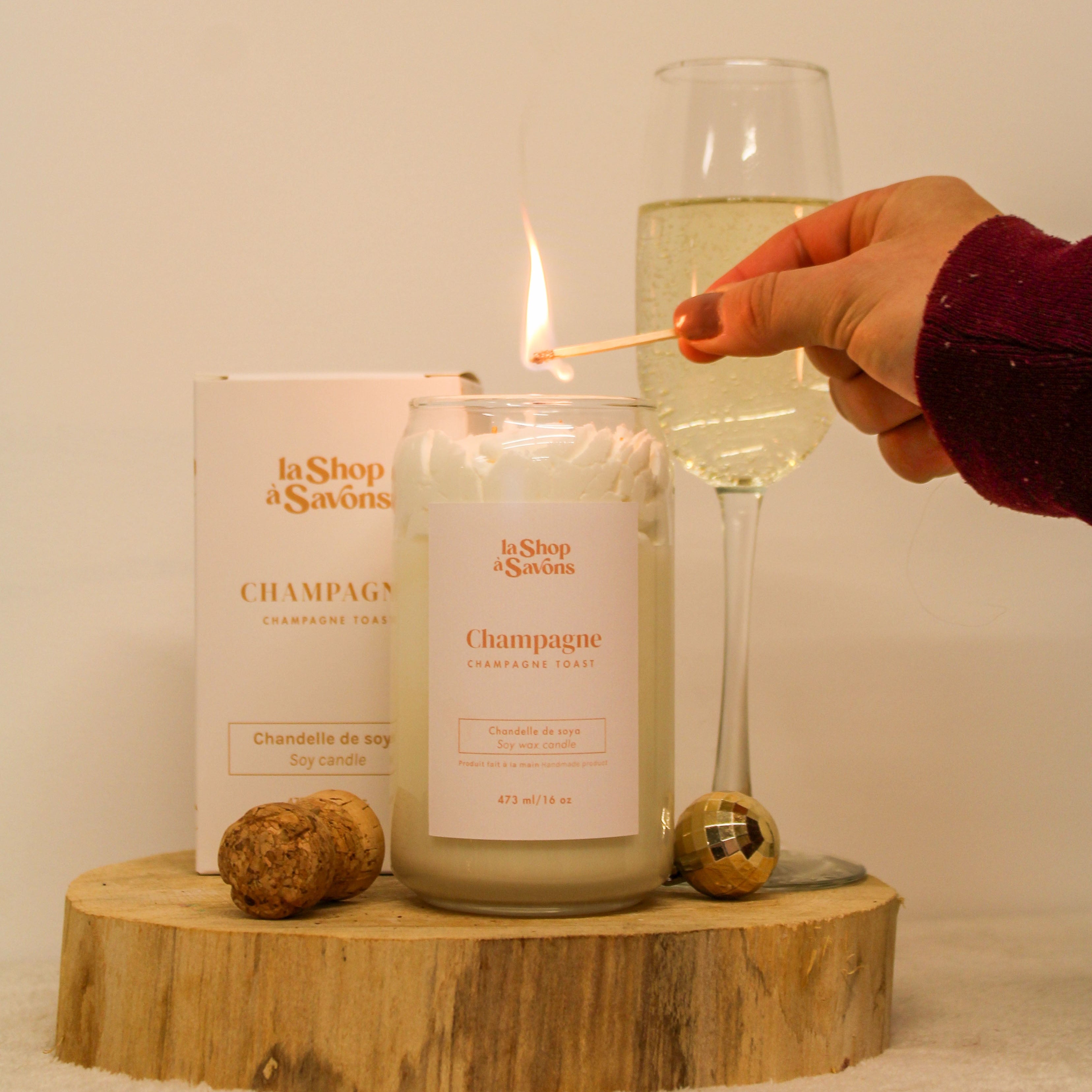 Candle labeled 'Champagne' being lit with a match, next to a glass of champagne and two corks on a wooden block.