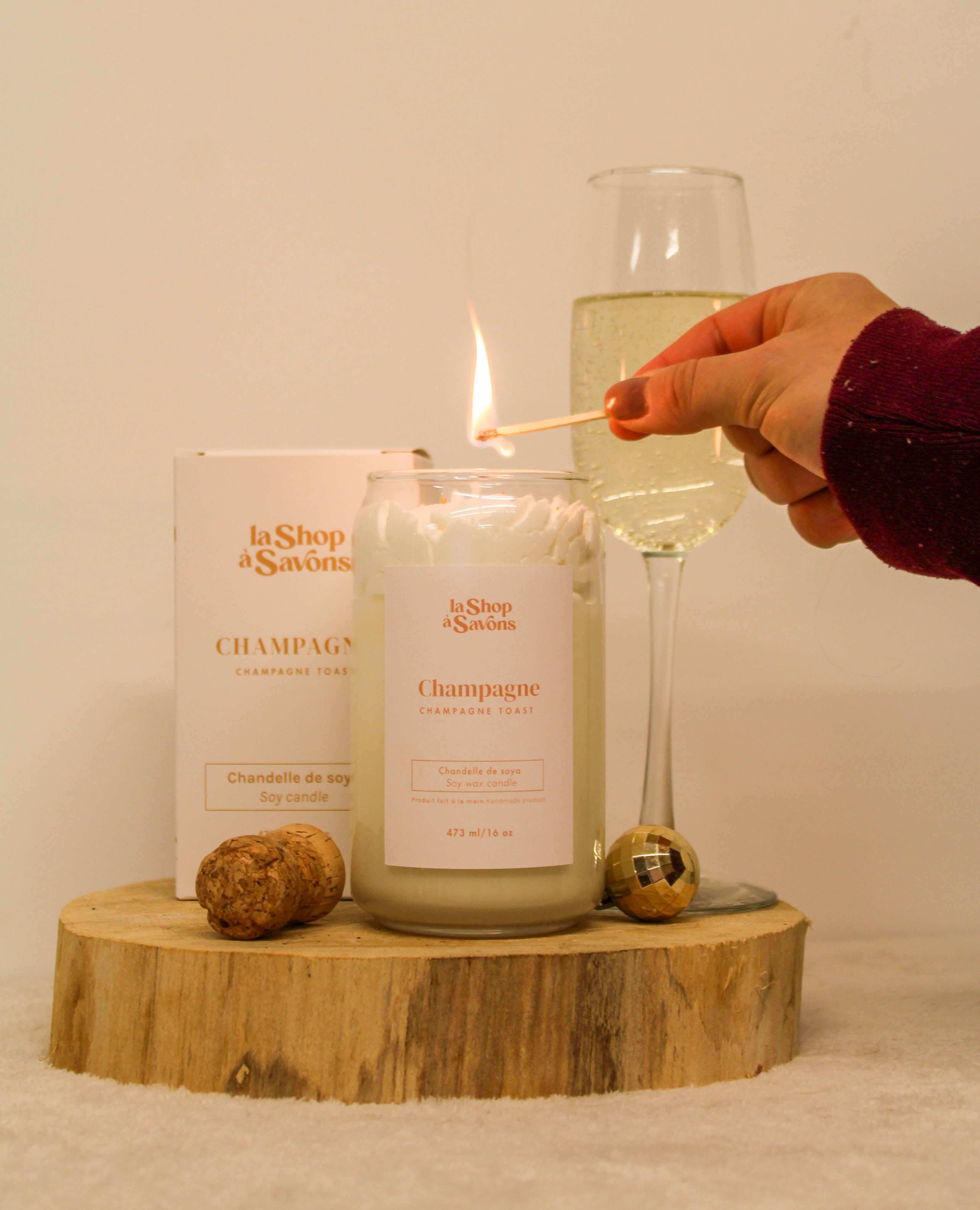 Candle labeled 'Champagne' being lit with a match, next to a glass of champagne and two corks on a wooden block.