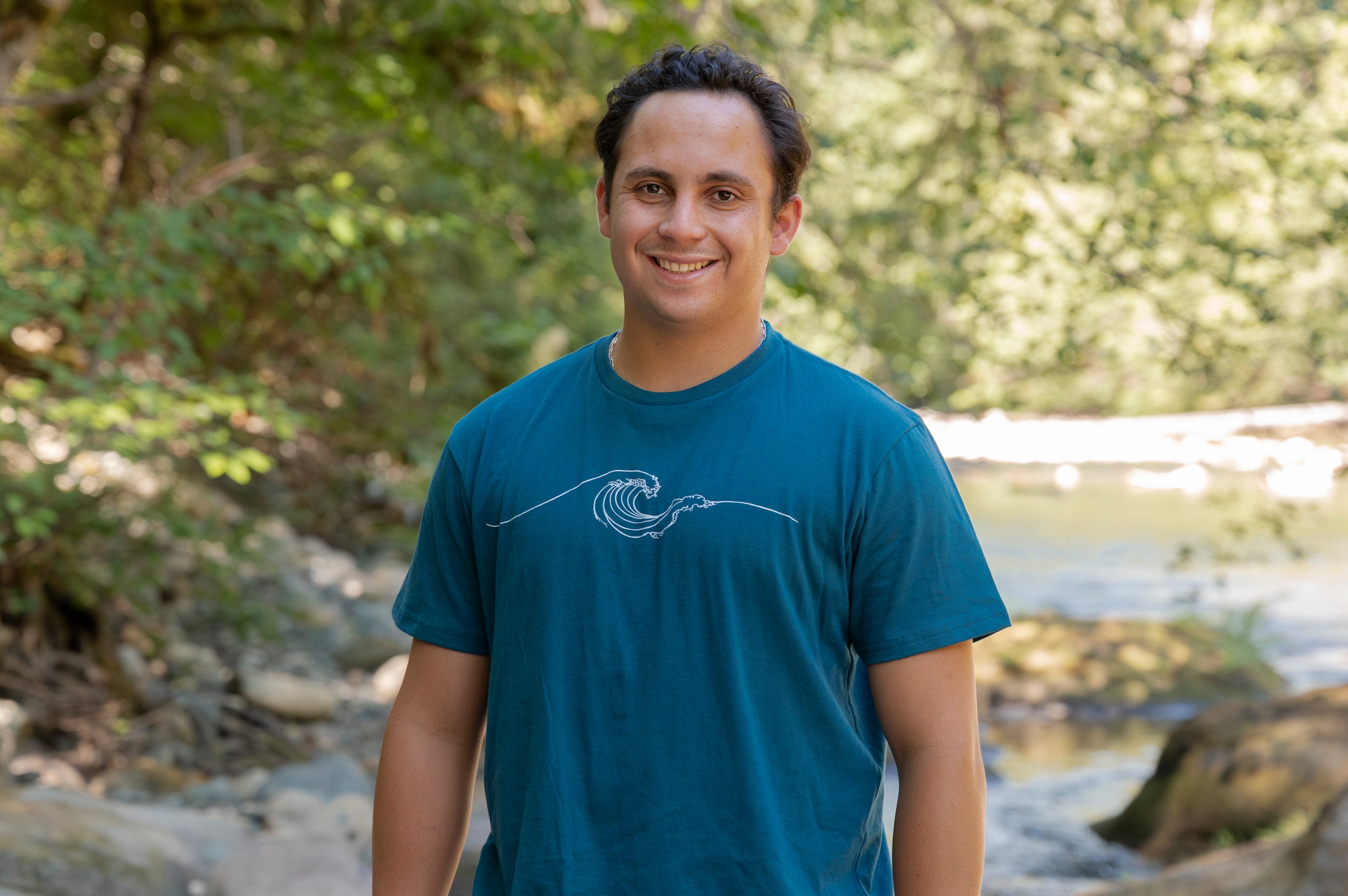 A man standing outdoors in a wooded area near a stream, wearing a teal blue t-shirt with a white wave graphic and beige shorts.