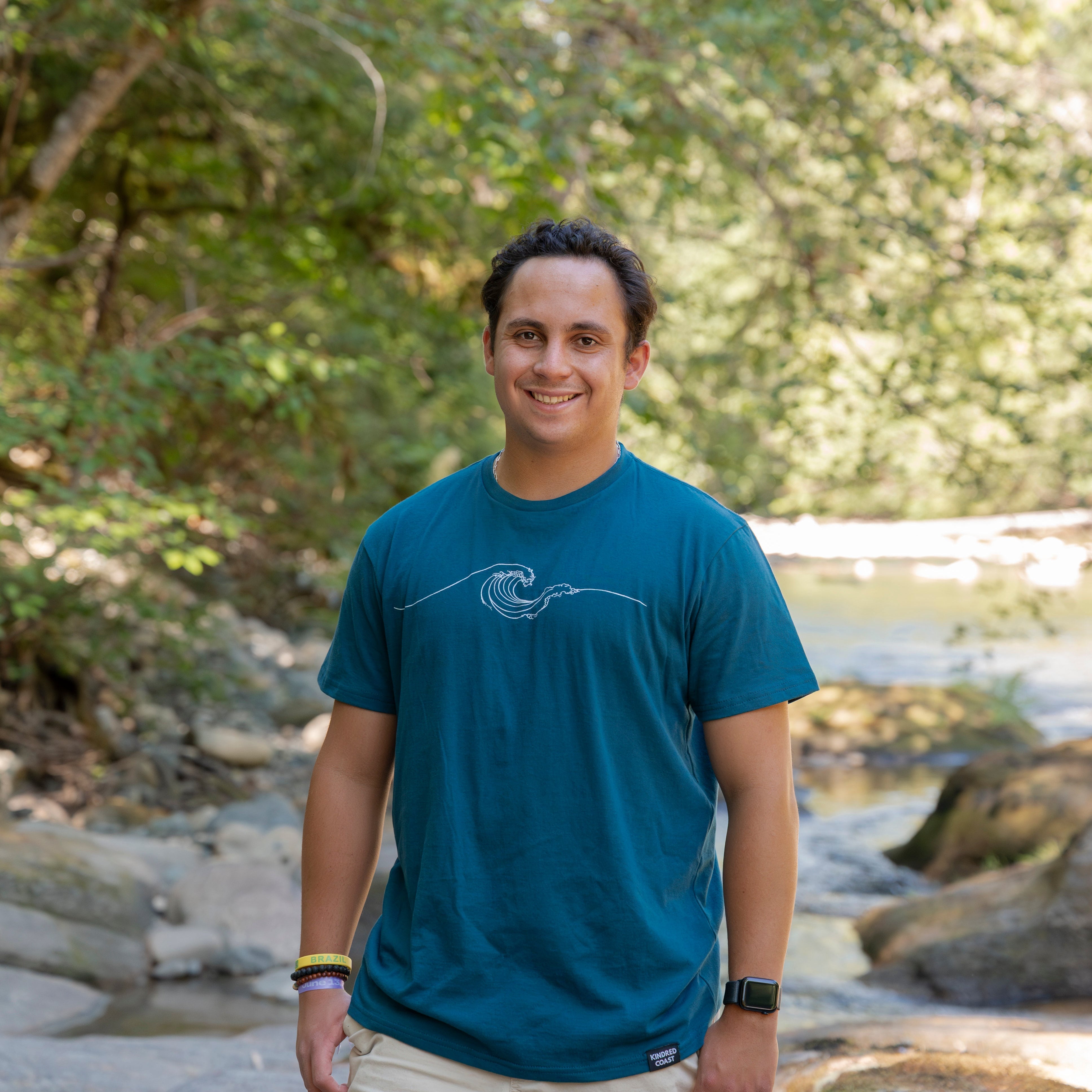A man standing outdoors in a wooded area near a stream, wearing a teal blue t-shirt with a white wave graphic and beige shorts.