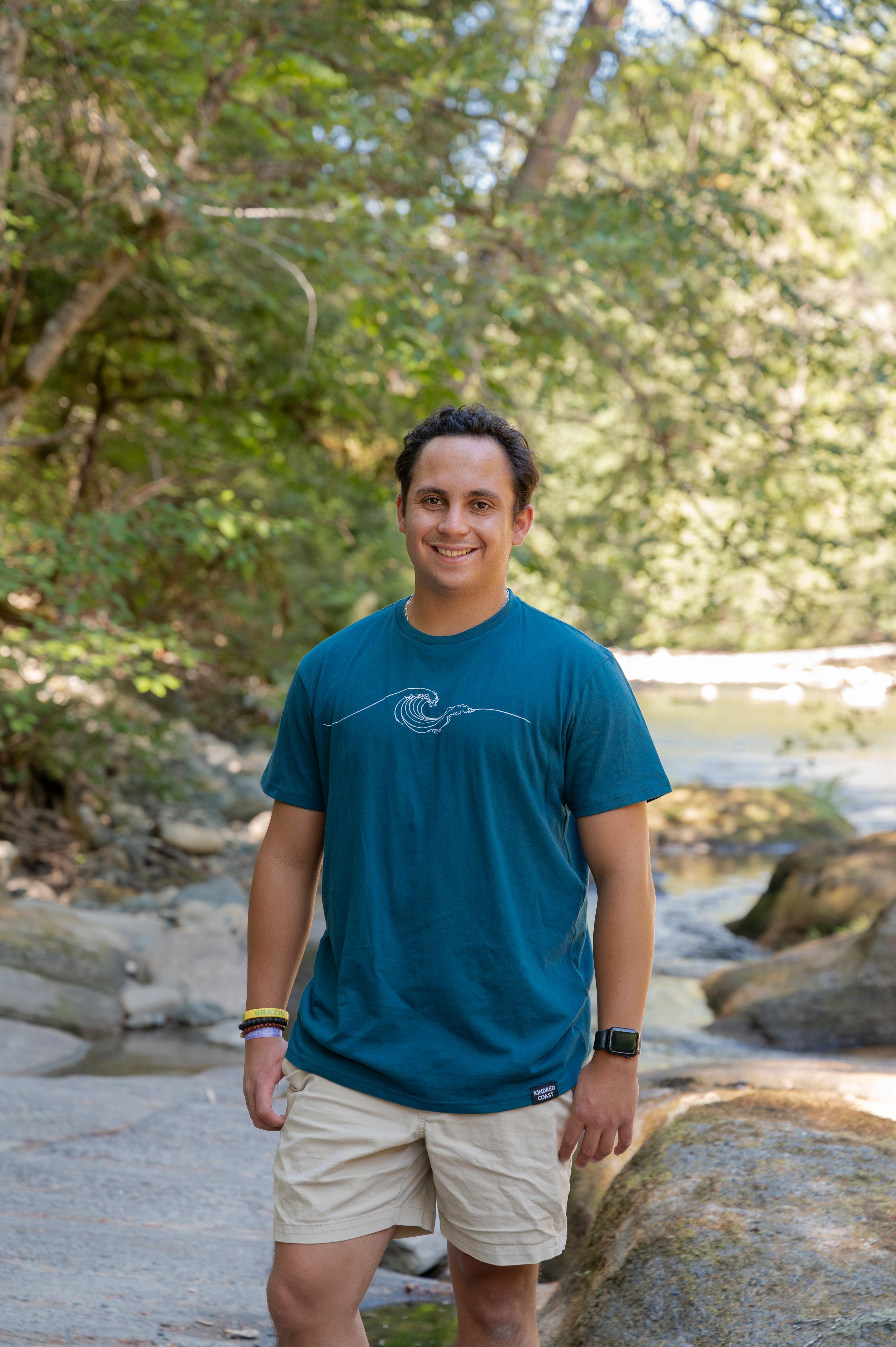 A man standing outdoors in a wooded area near a stream, wearing a teal blue t-shirt with a white wave graphic and beige shorts.