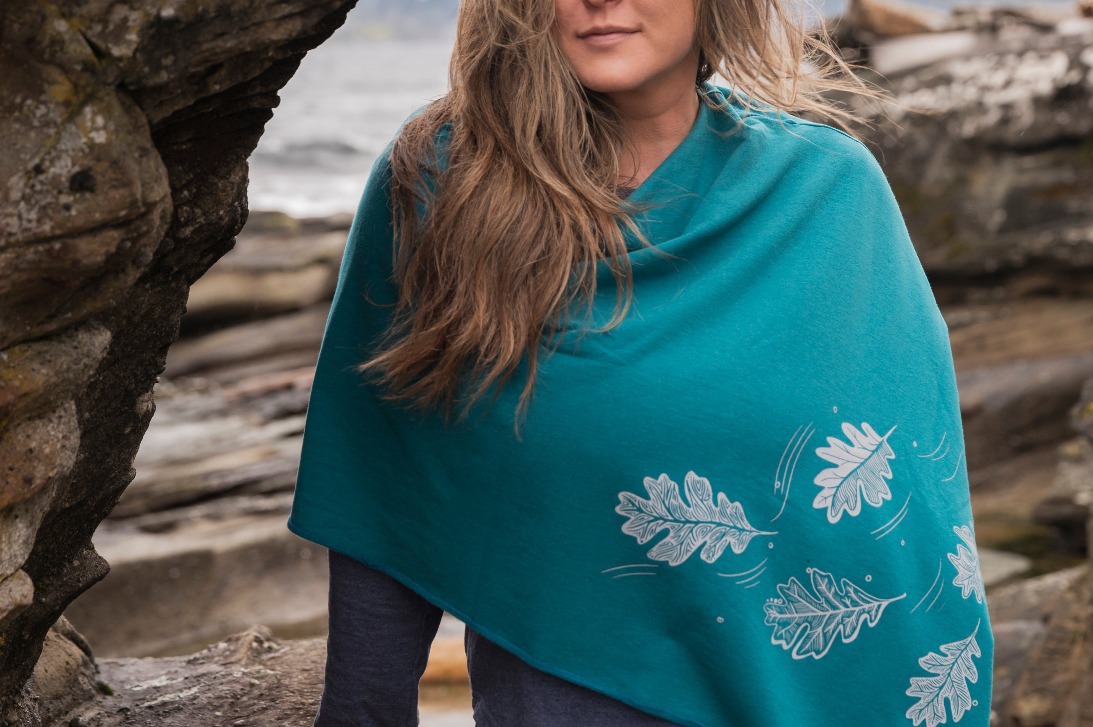 Woman wearing a teal poncho with leaf patterns by a rocky coastal area