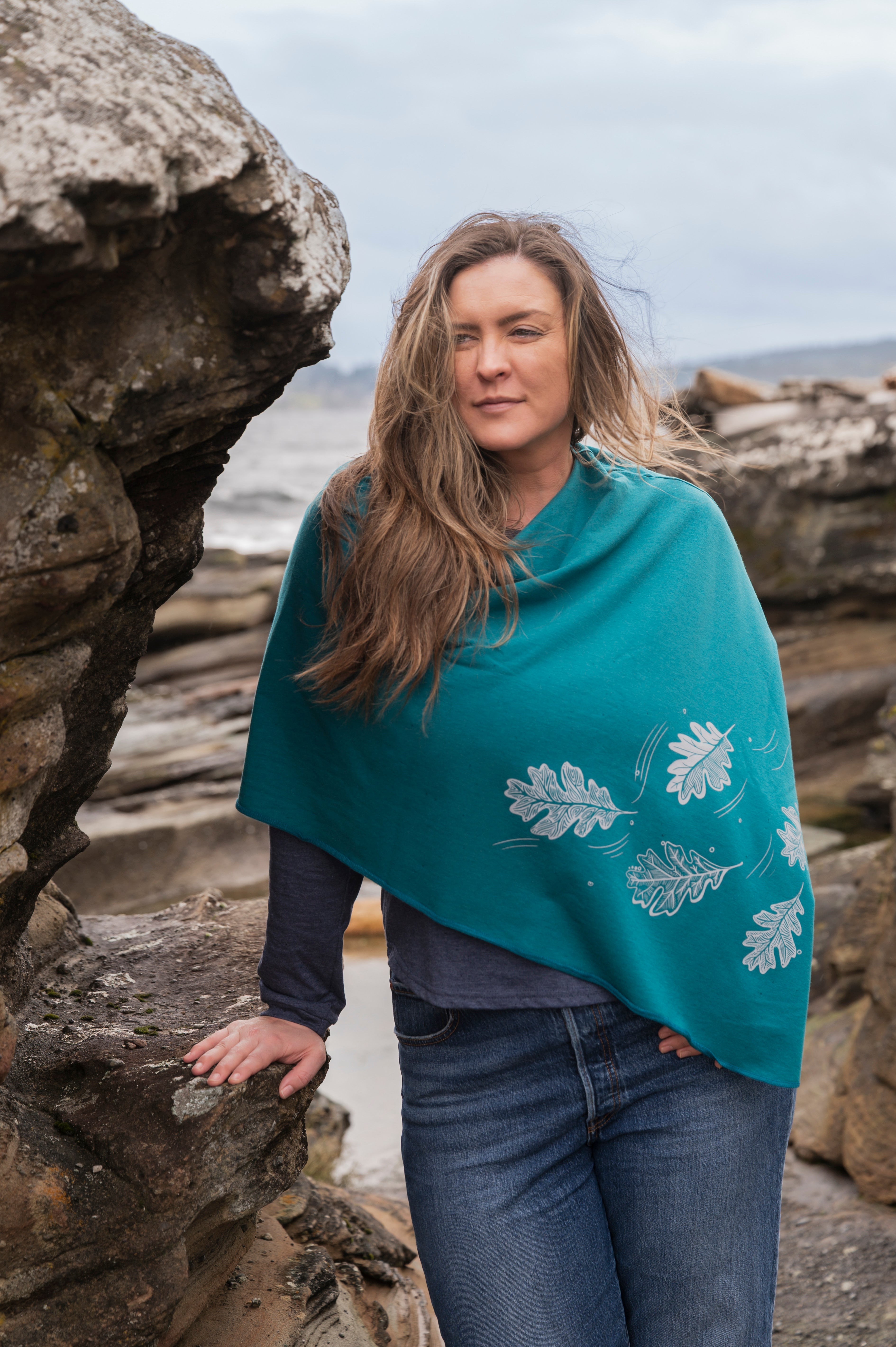 Woman wearing a teal poncho with leaf patterns by a rocky coastal area