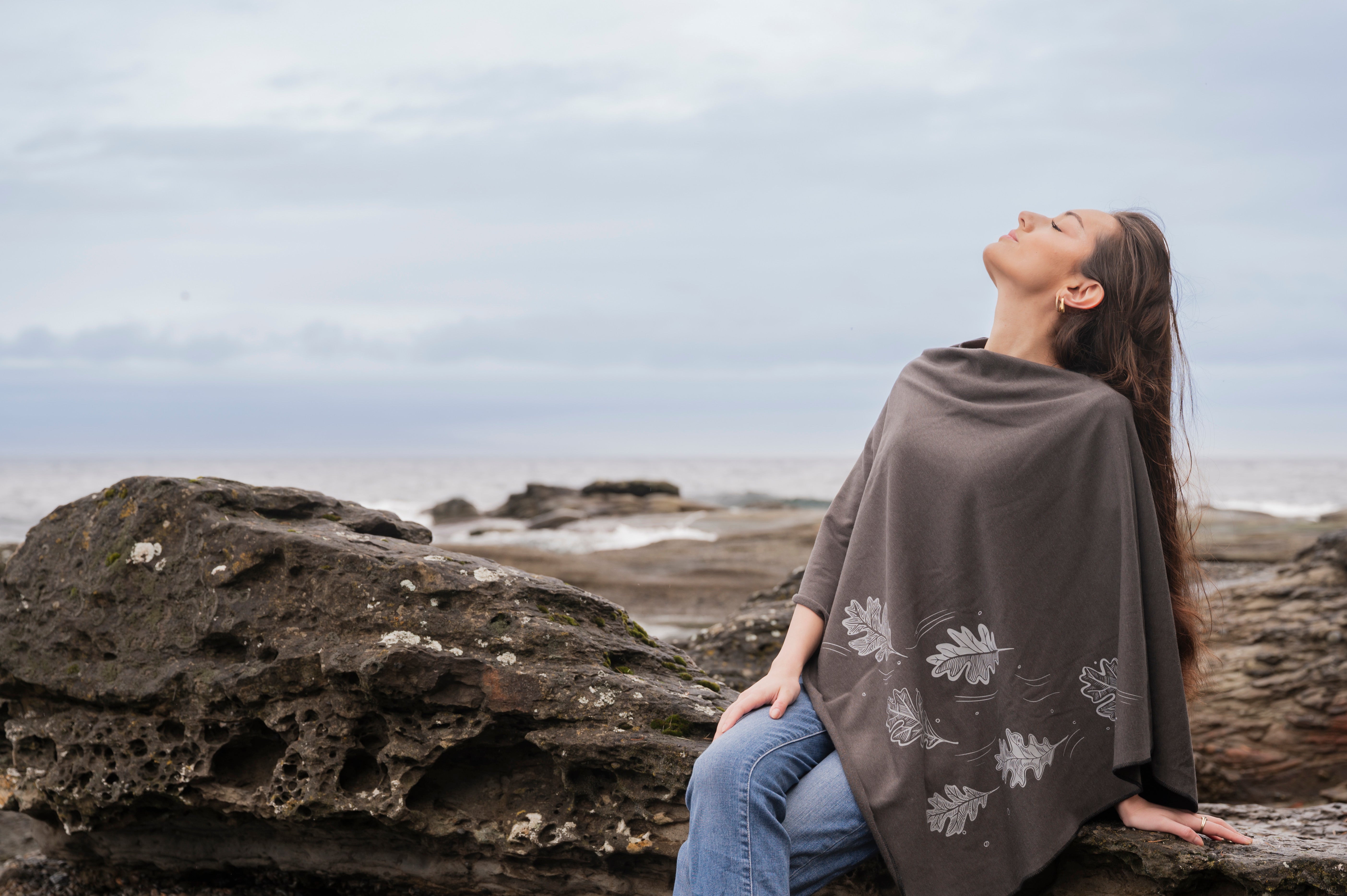 Woman sitting on a rock by the ocean with a patterned blanket draped over her shoulders.