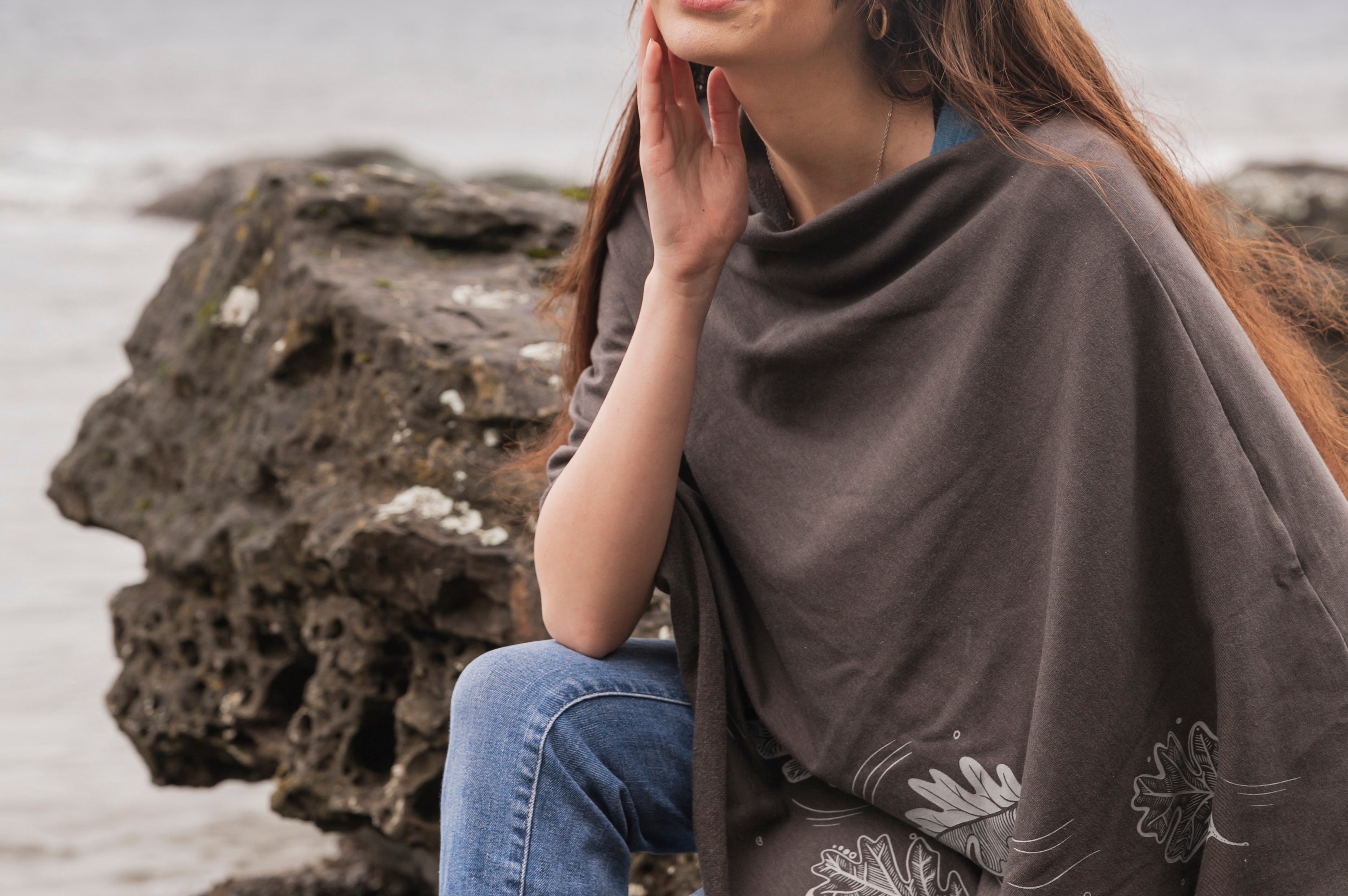 Woman sitting on a rock by the sea wearing a brown poncho with floral patterns.