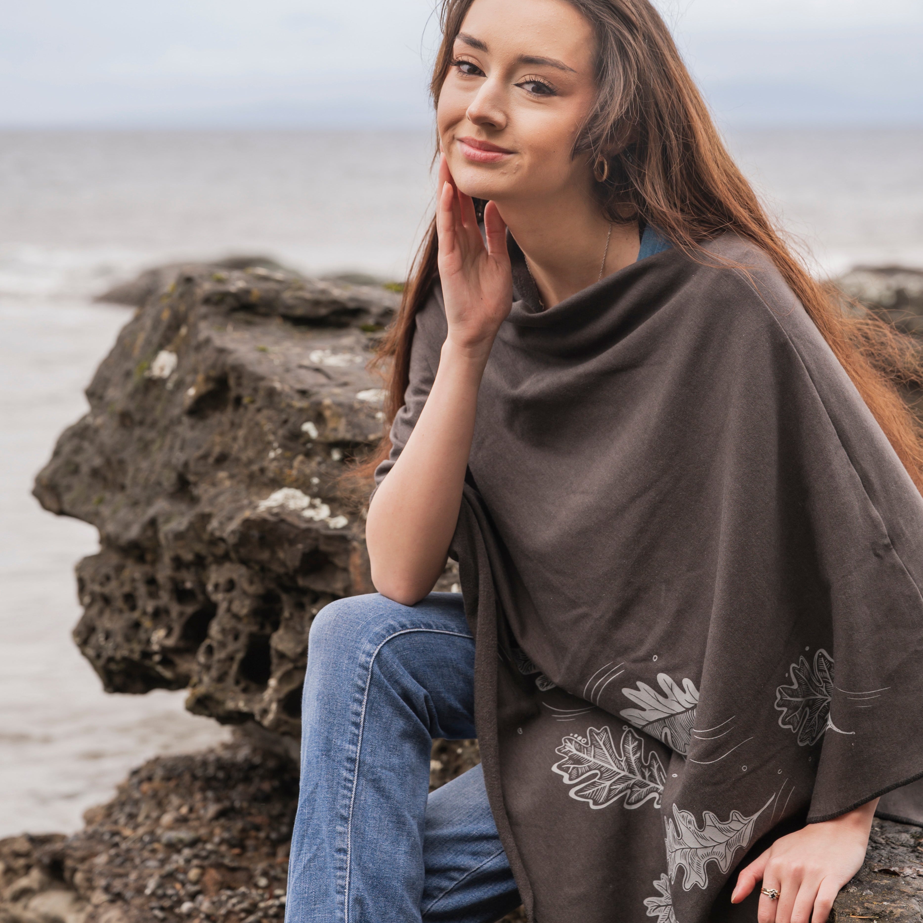 Woman sitting on a rock by the sea wearing a brown poncho with floral patterns.
